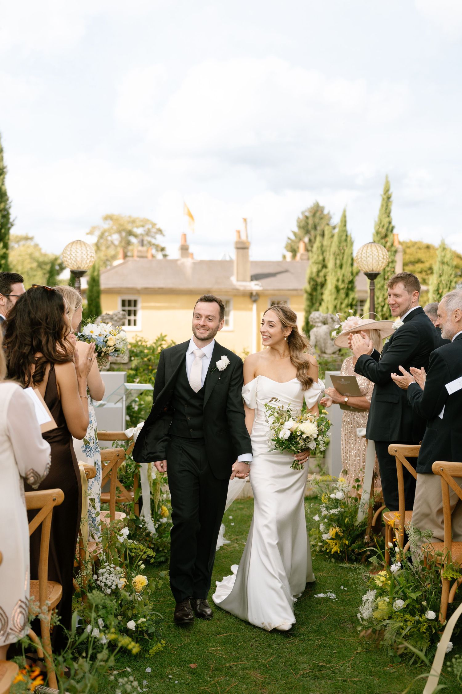 Wedding party and guests mingle on lawn after outdoor ceremony with cypress trees in background.