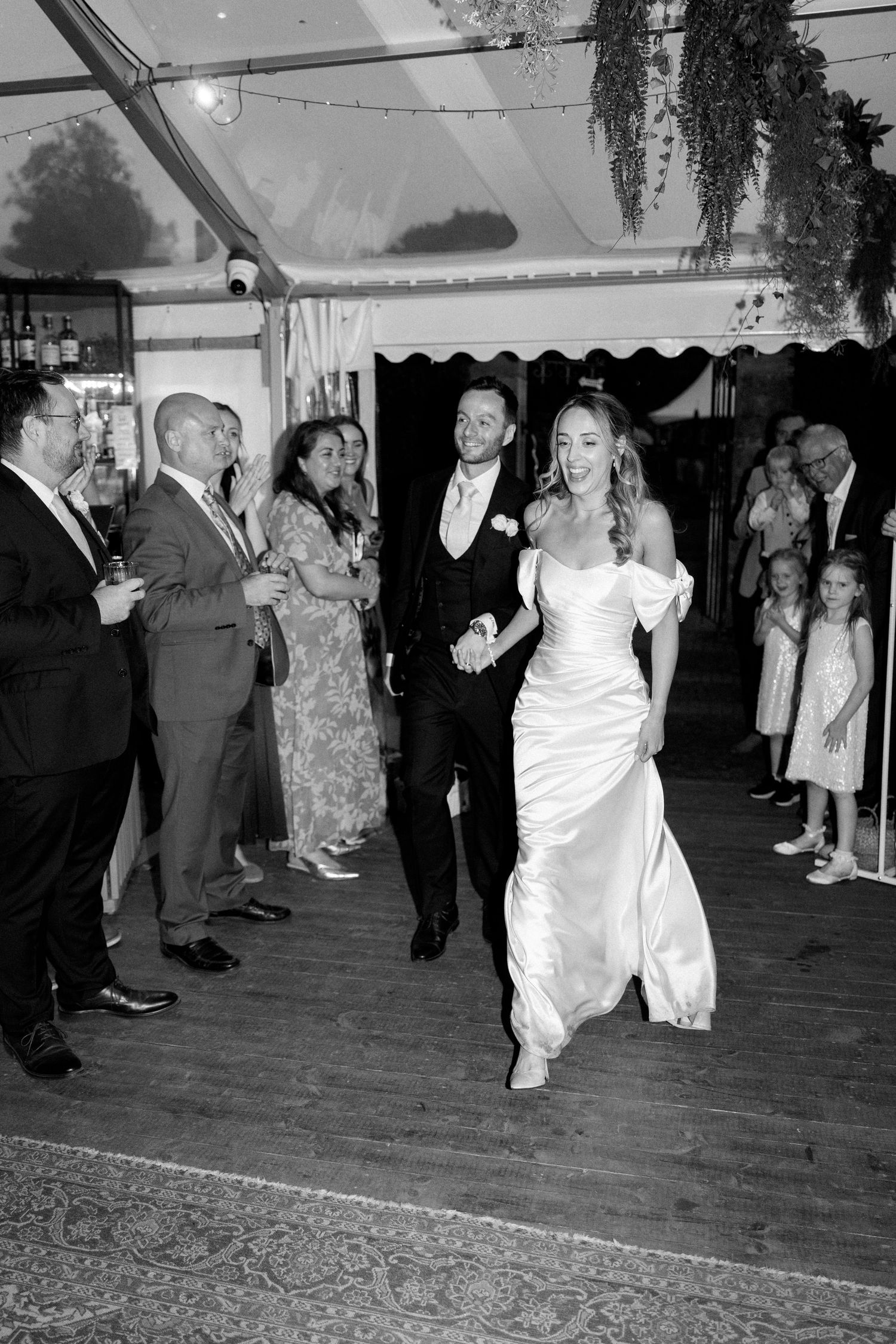 A black and white photo captures a wedding couple's entrance at their reception with guests looking on.