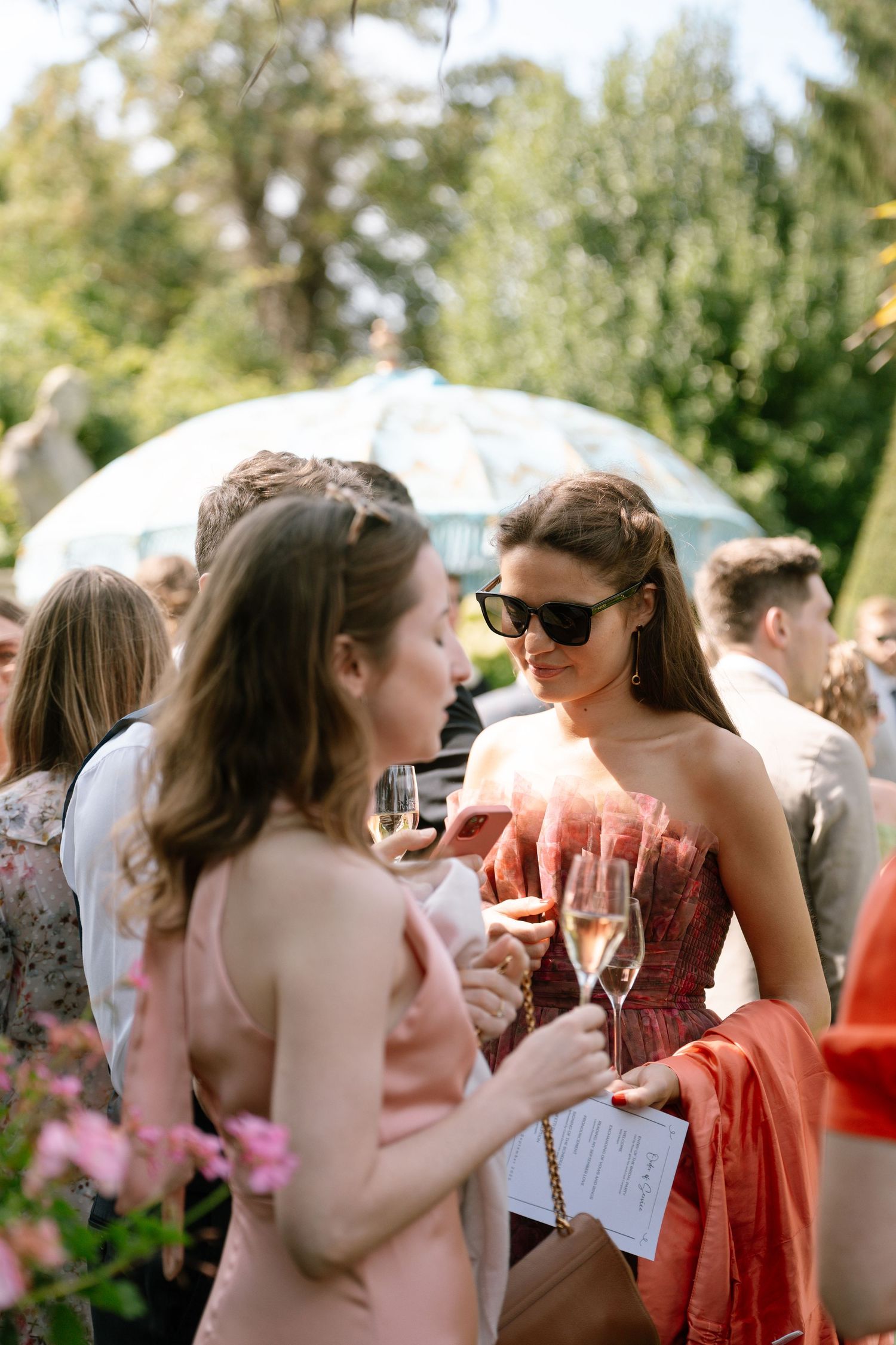 Wedding guests in colorful dresses chat and enjoy champagne during an outdoor reception.