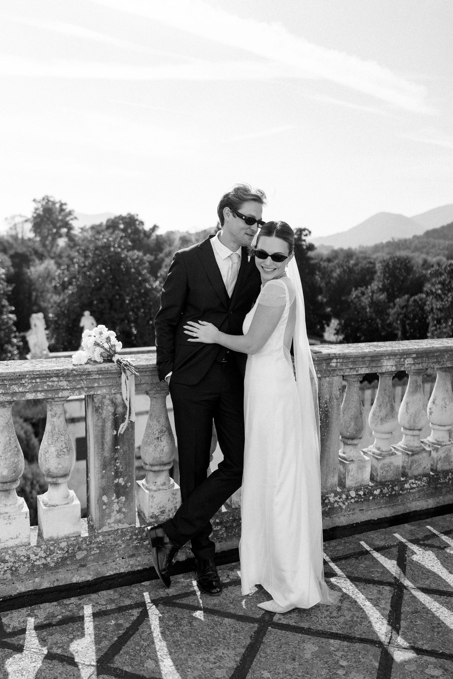 A stylish couple in sunglasses pose together on a historic terrace with mountain scenery.