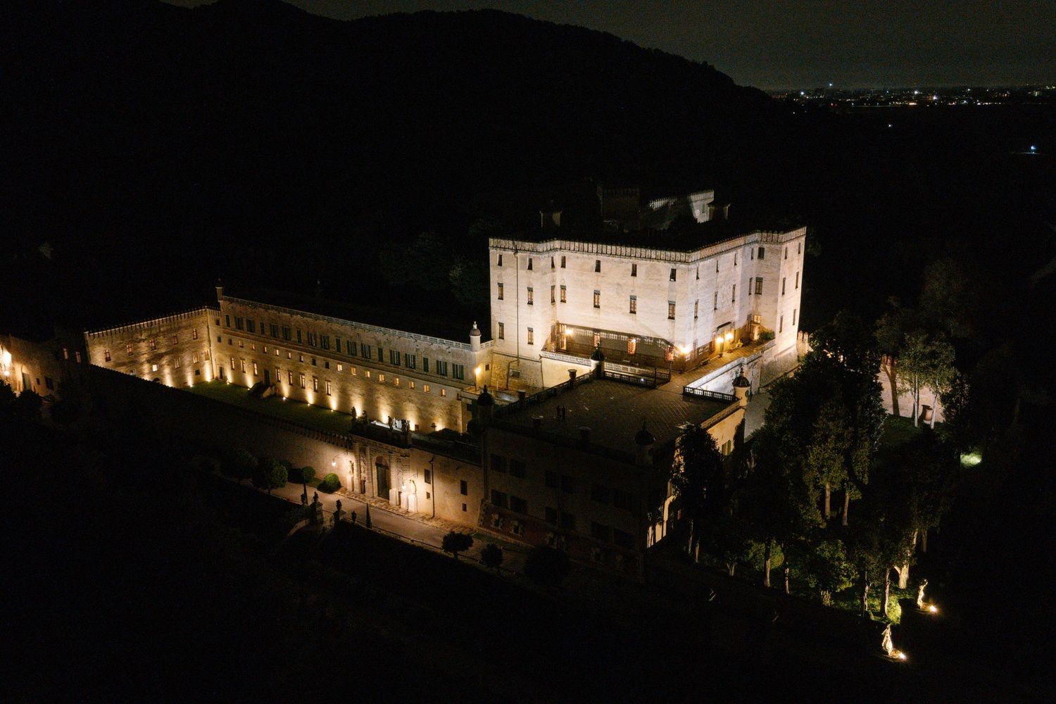 Night view of illuminated castle buildings along a hillside.