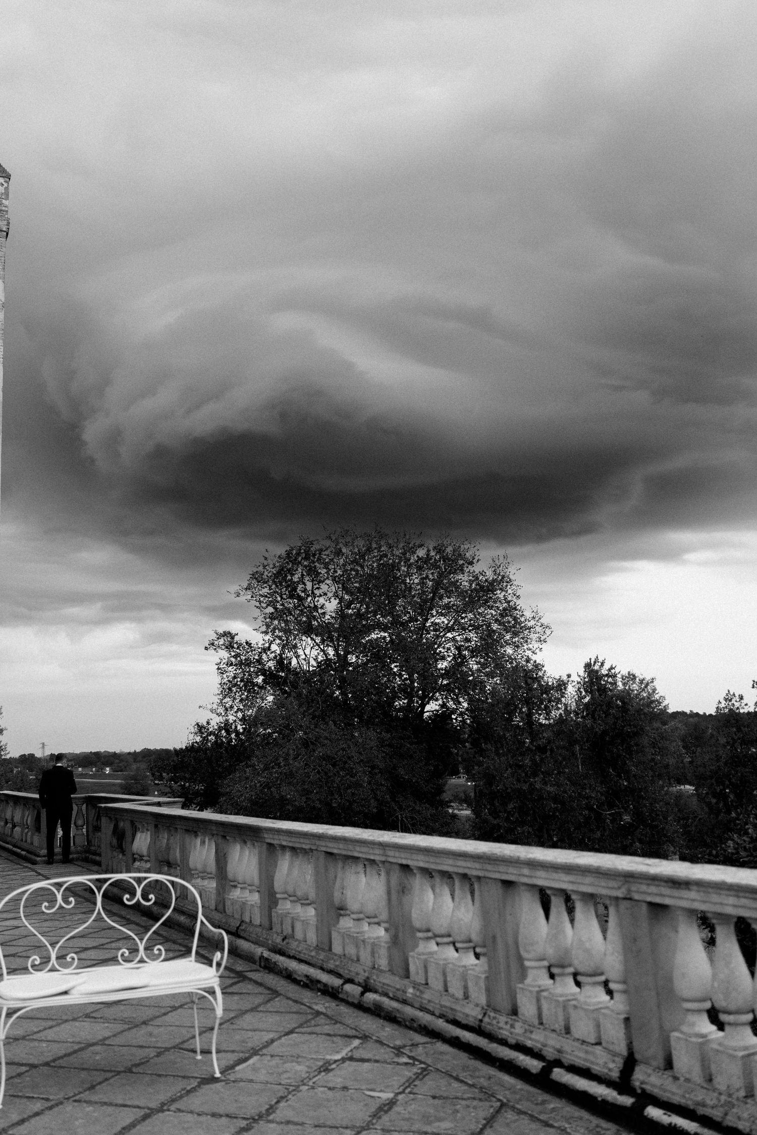 Dramatic storm clouds gather over stone balcony with white metal chairs.