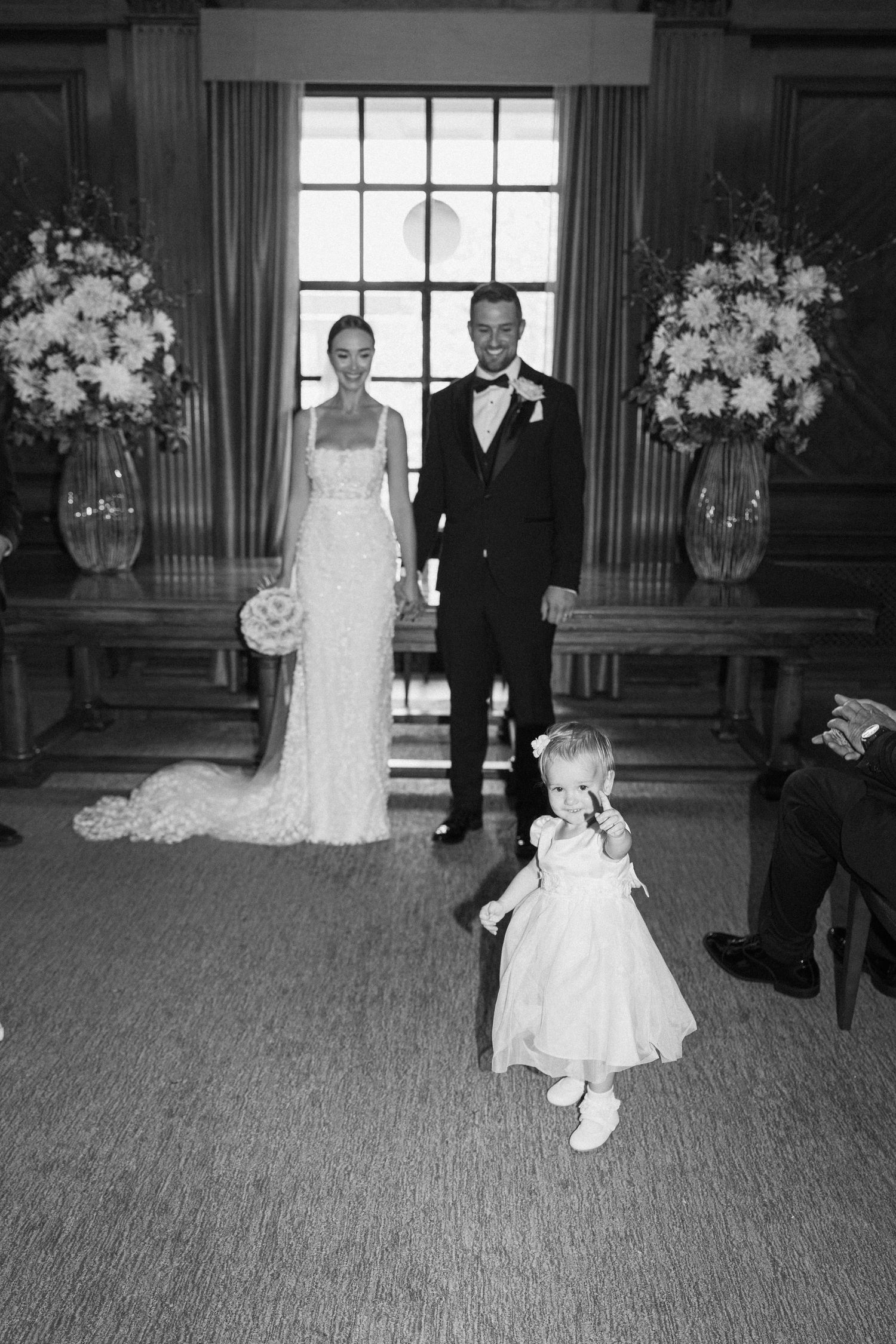 Black and white wedding photos showing couple walking across carpeted room with floral arrangements.