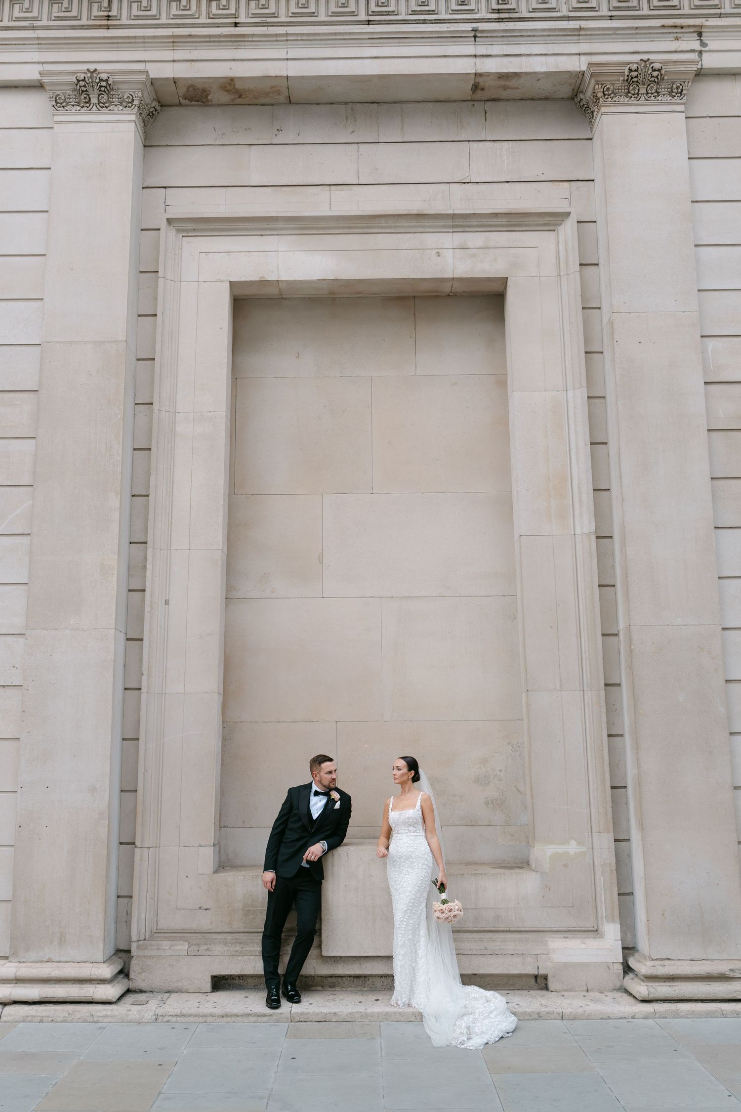 Wedding couple poses against a grand classical stone archway in formal attire.