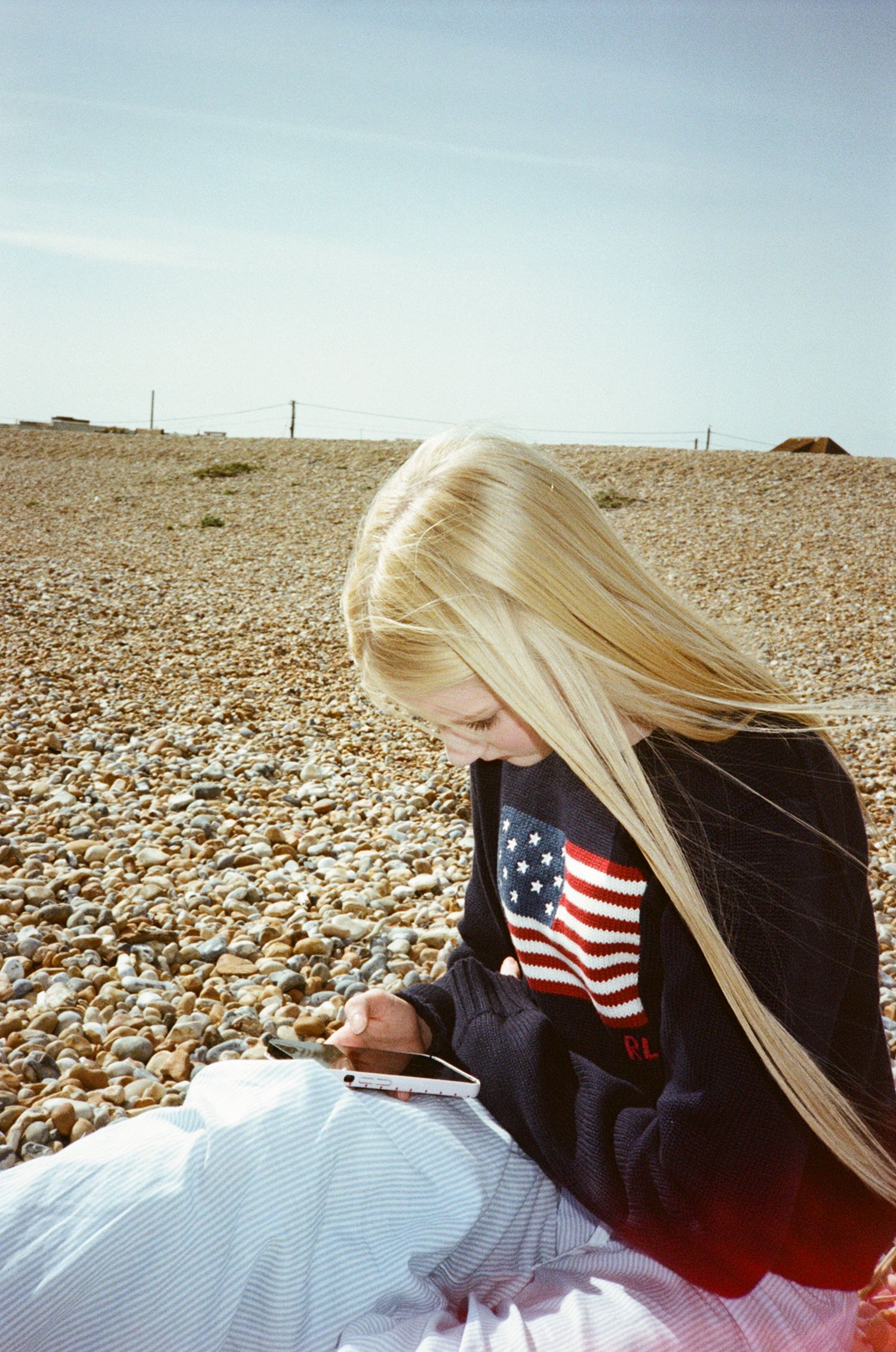 Blonde girl in American flag sweatshirt sitting on pebble beach looking at her phone on a sunny day.