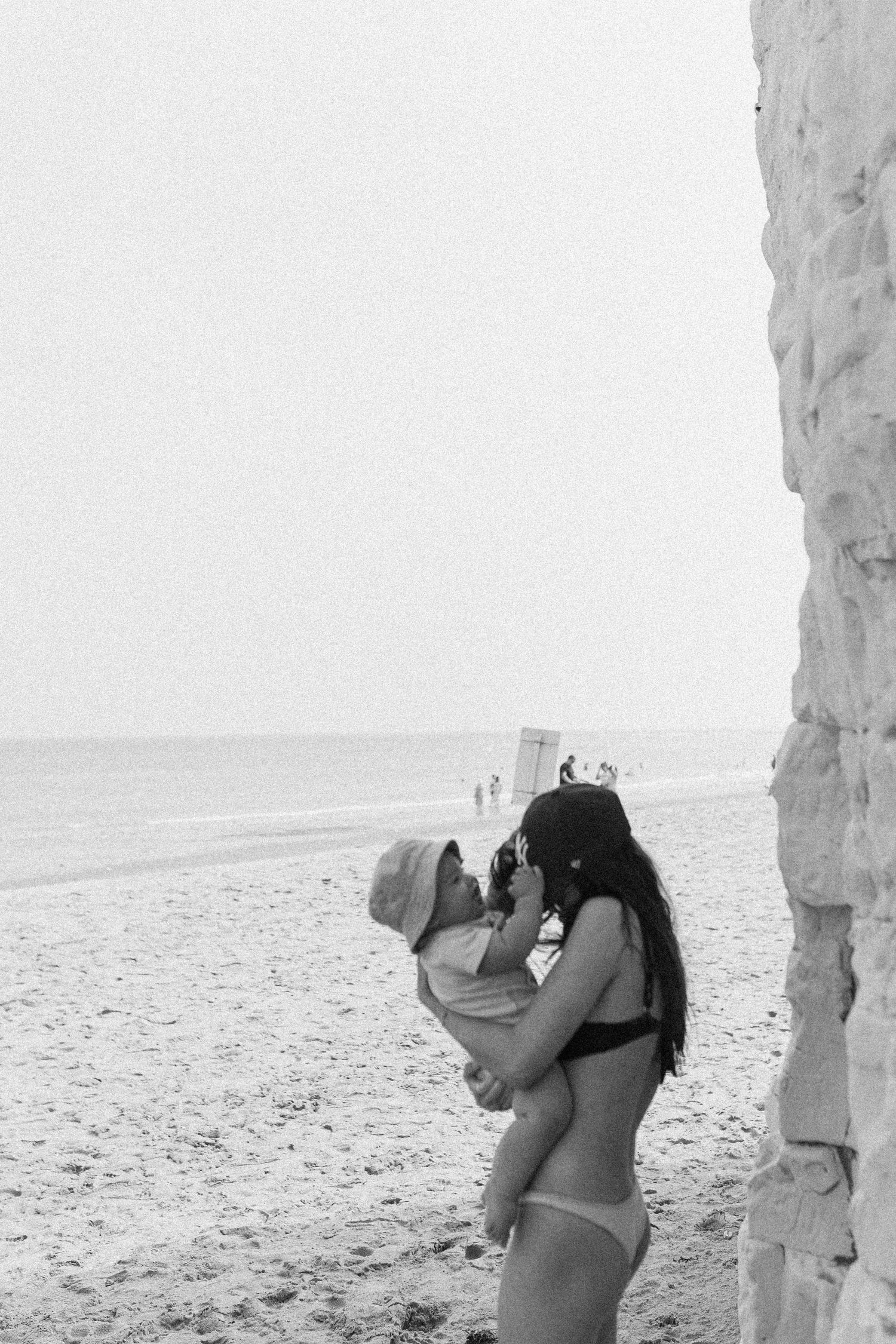 Black and white photo of a woman in a bikini holding a baby on a sandy beach near a stone wall.