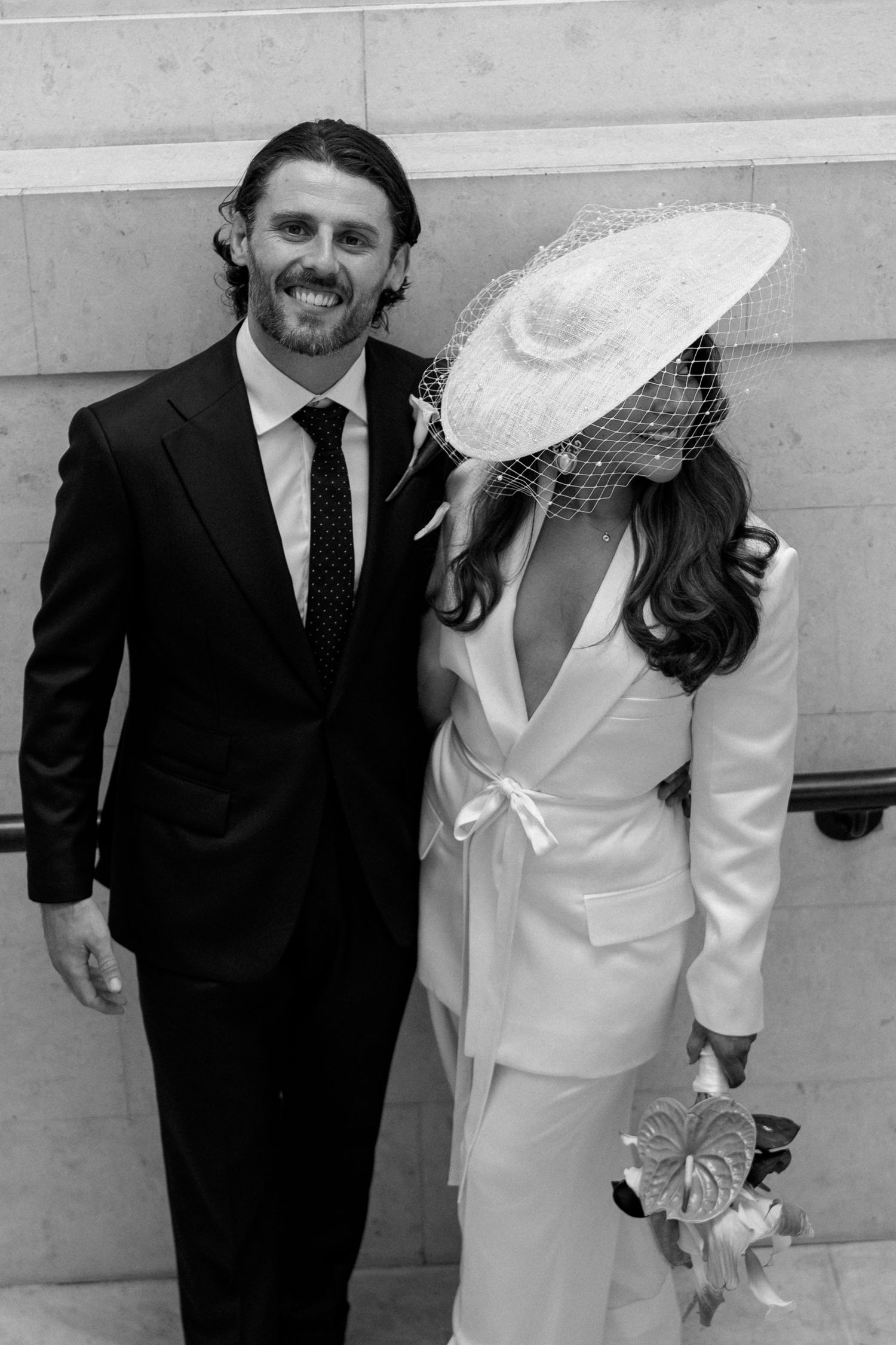 Black and white photo of a smiling groom in a dark suit beside a bride in a white suit and wide-brim veil hat holding a bouquet.