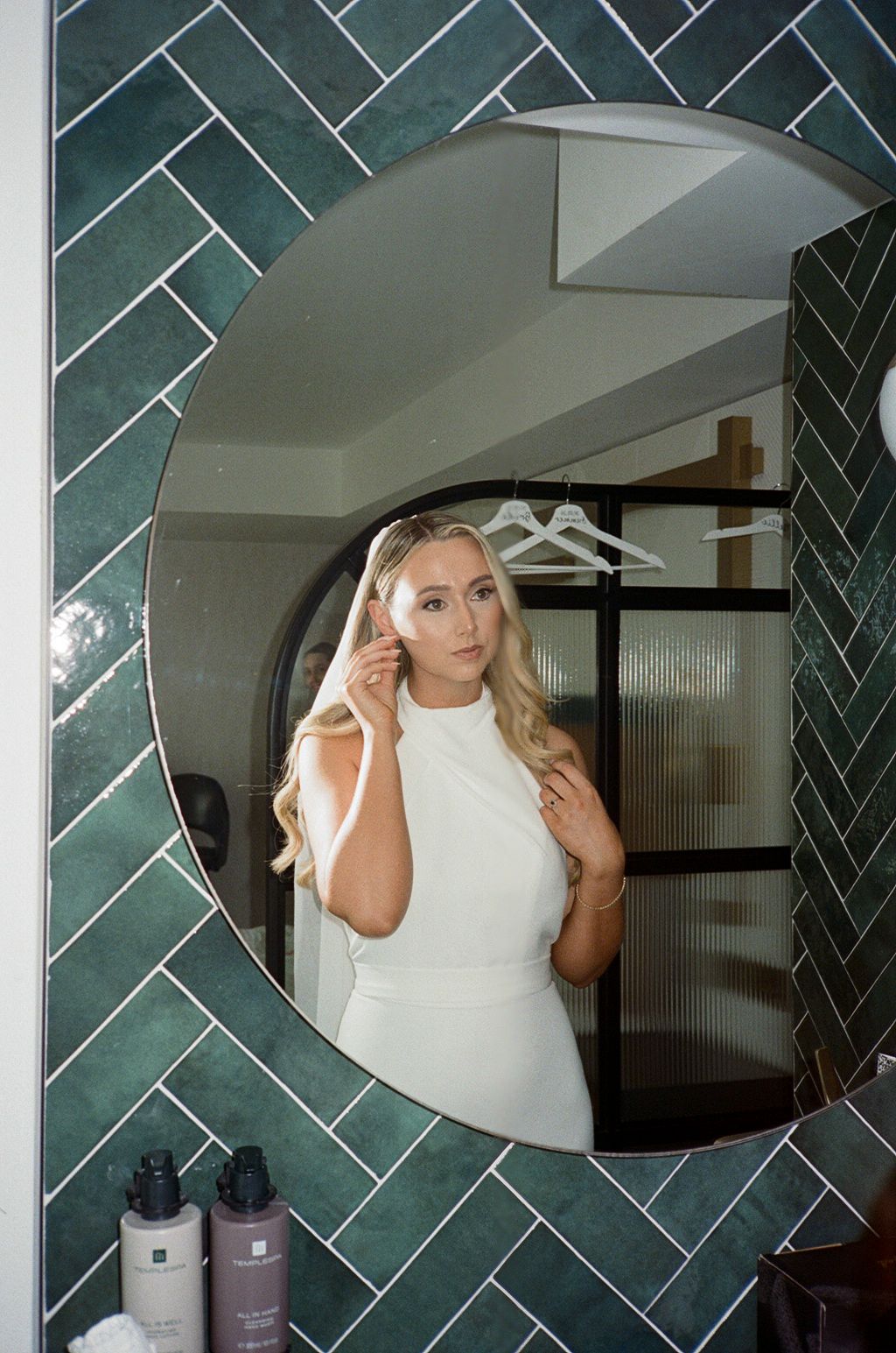 Blonde woman in white dress adjusting earring reflected in round mirror with green herringbone tile bathroom wall.