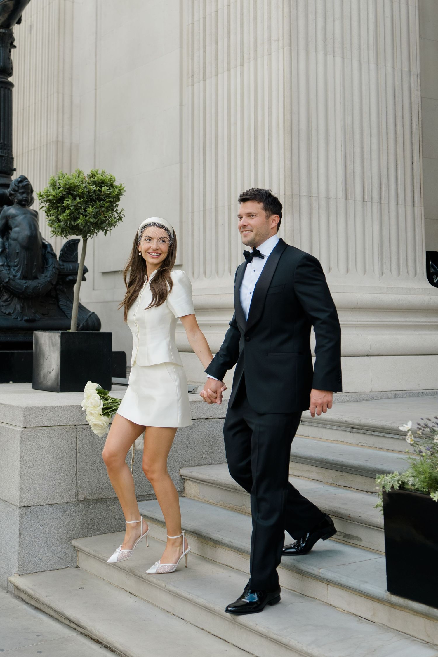 A bride in a white mini dress and groom in a black tuxedo hold hands while descending stone steps at their wedding.
