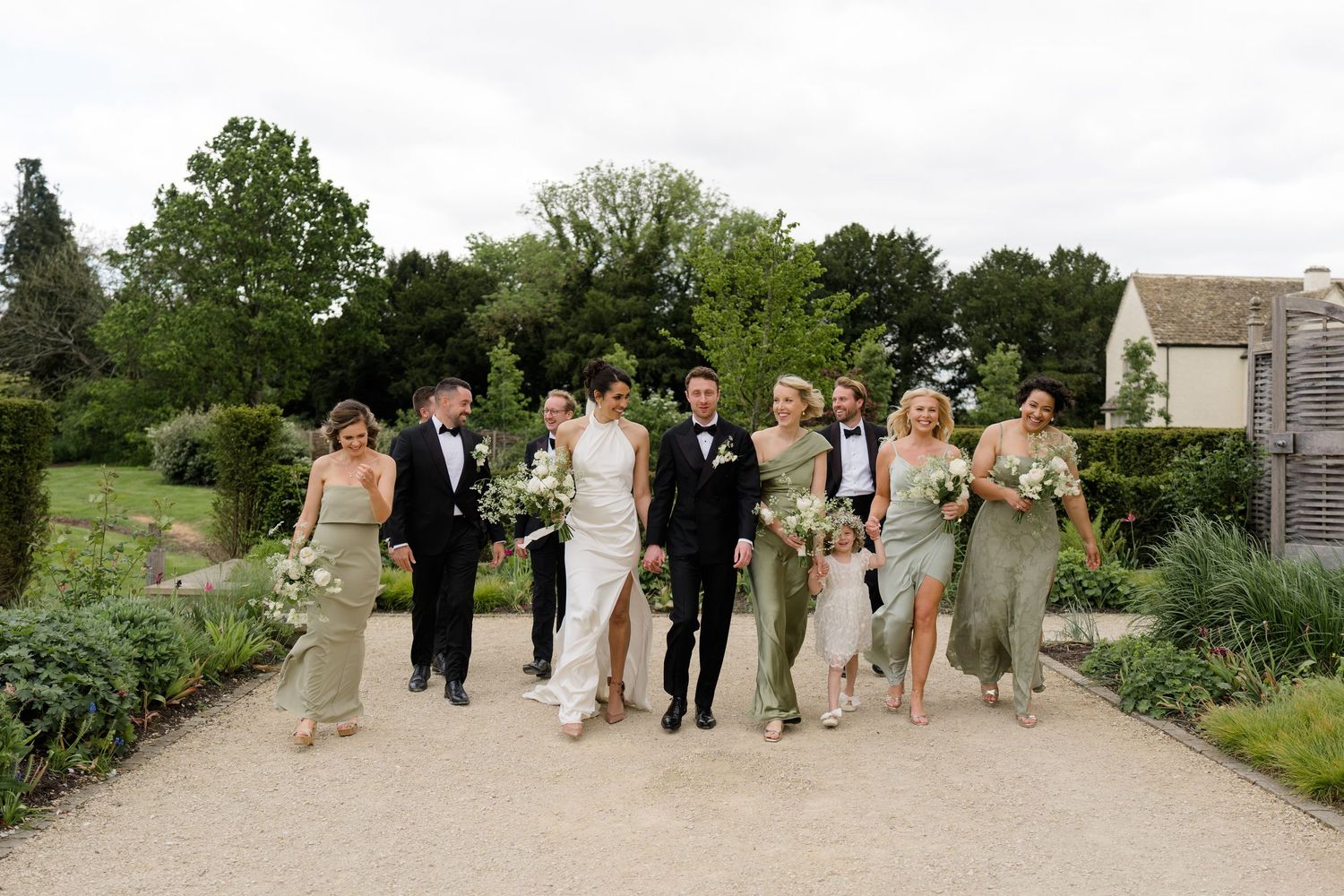 Wedding party walks together in garden, bridesmaids in sage green gowns with groomsmen in black tuxedos.
