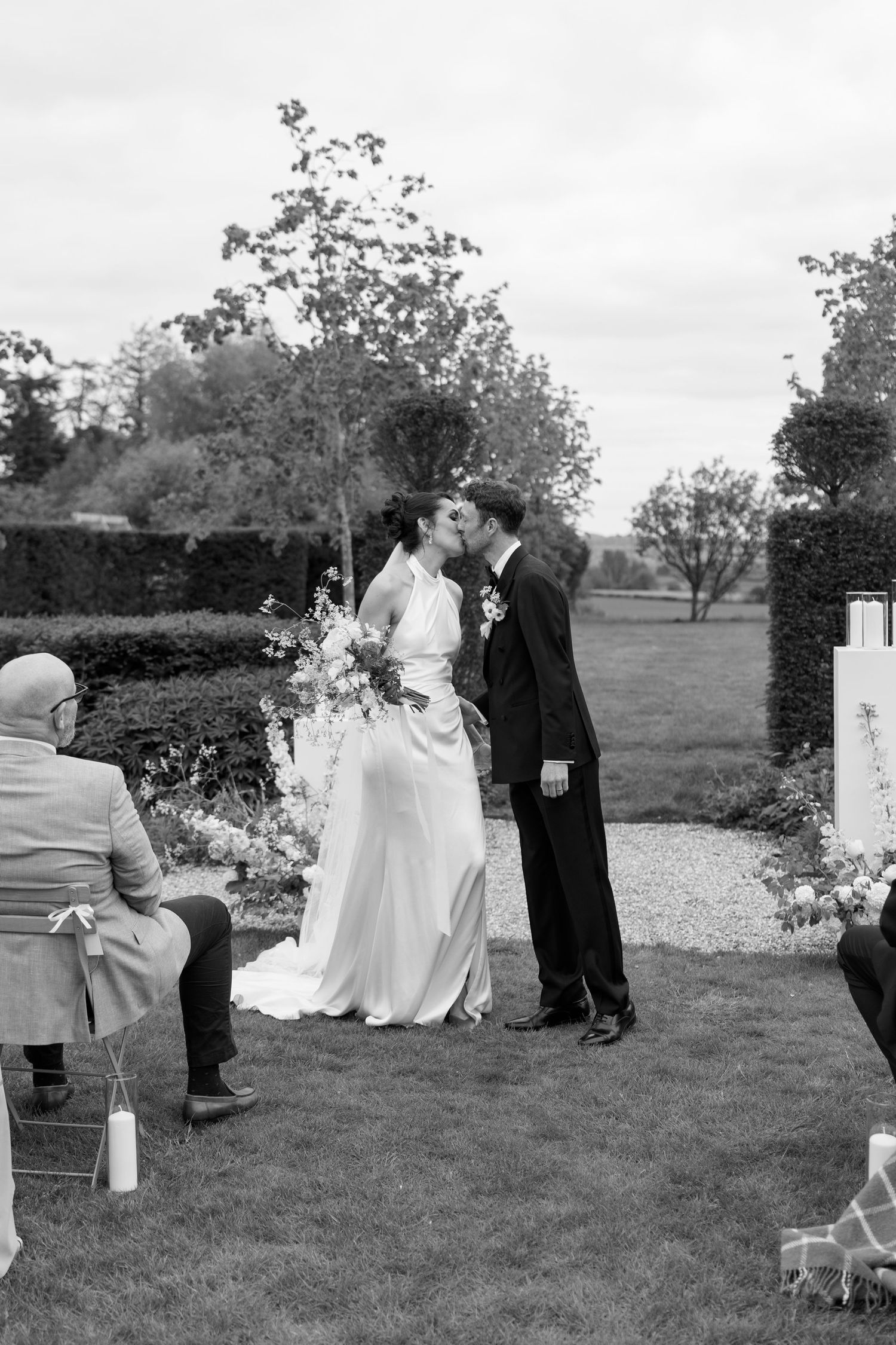 Black and white photo of newlyweds sharing first kiss at outdoor garden wedding ceremony.