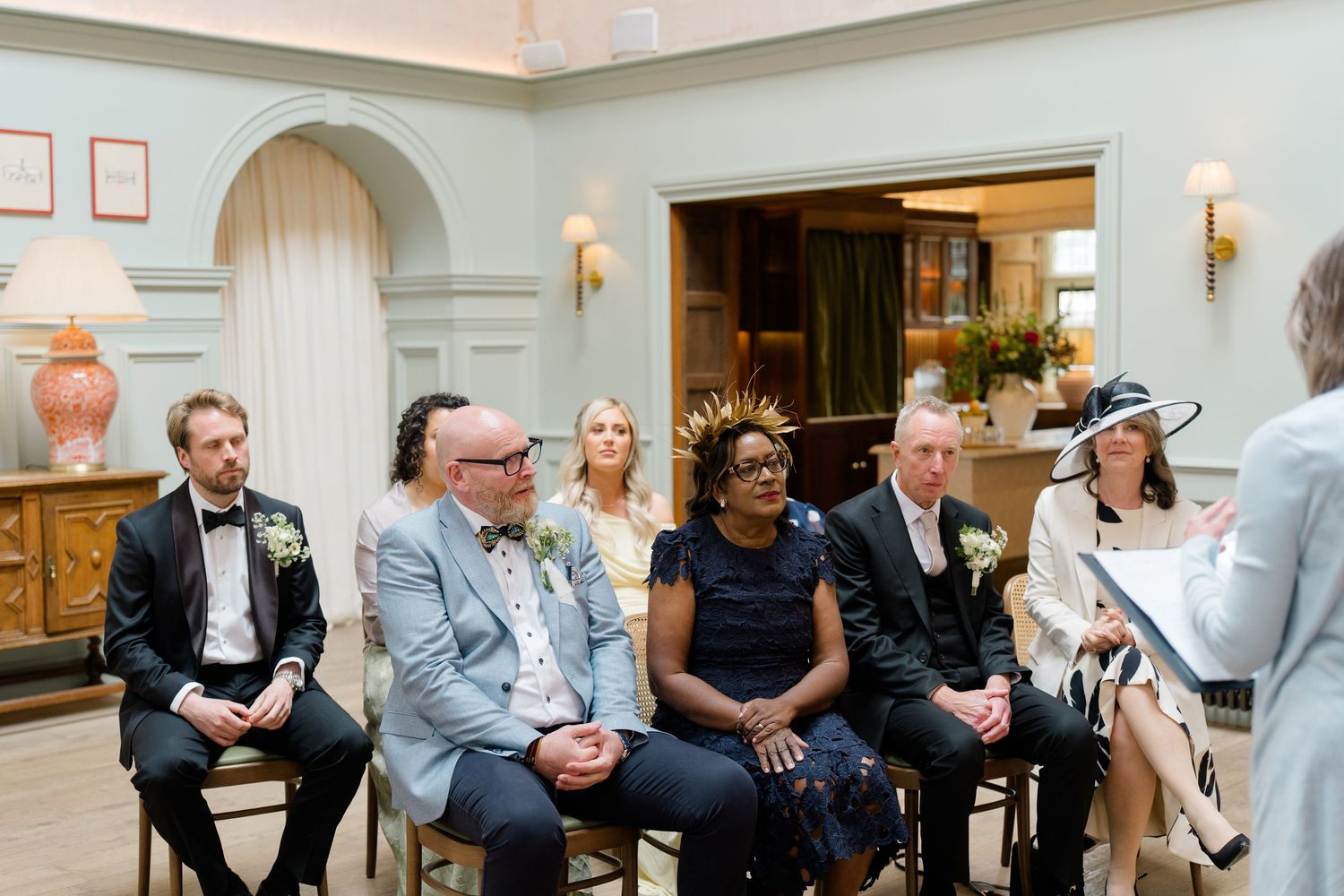 Wedding guests seated indoors watch ceremony as officiant reads from notes at altar.