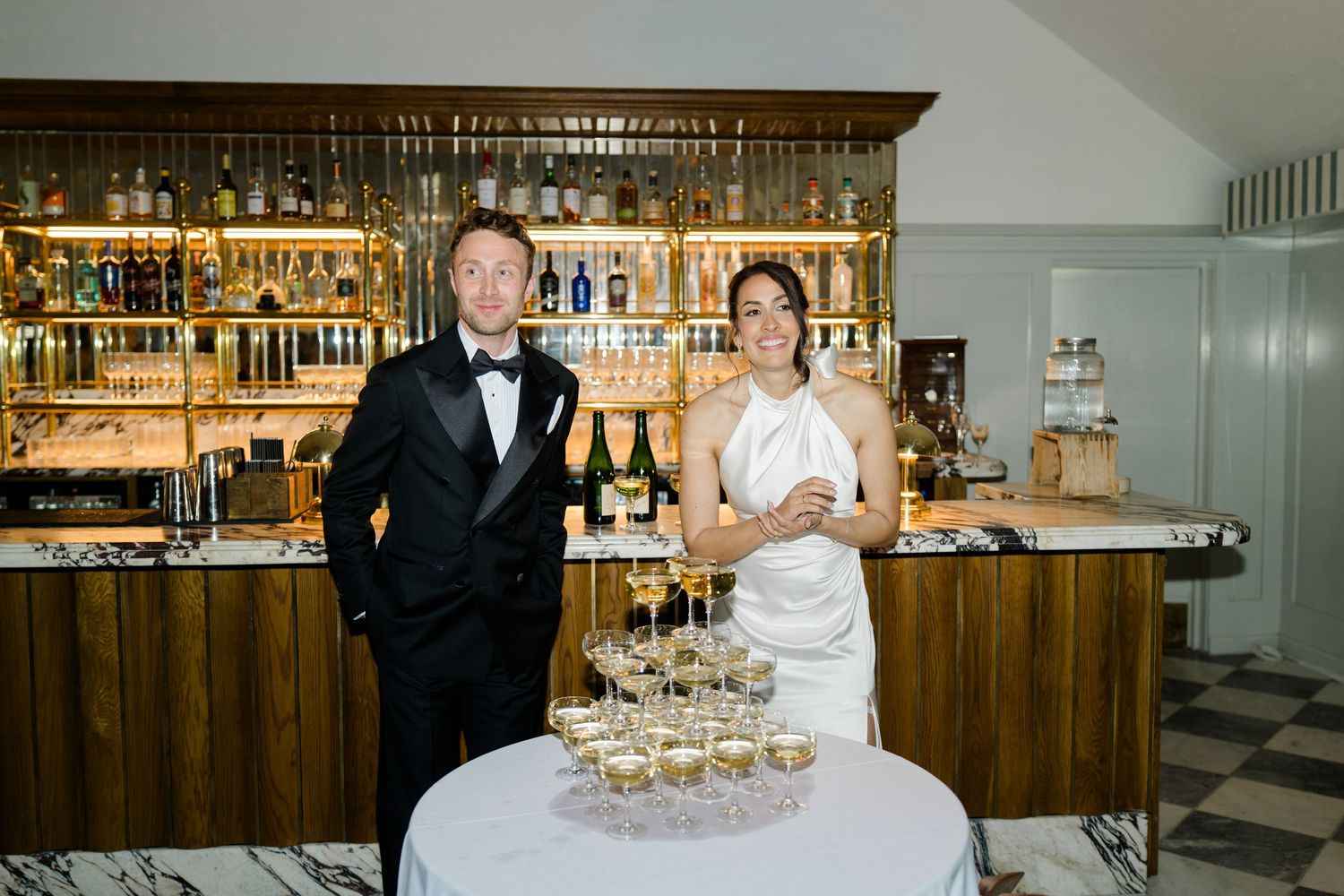 Bride and groom pose beside a champagne tower at a bar during their elegant wedding reception.