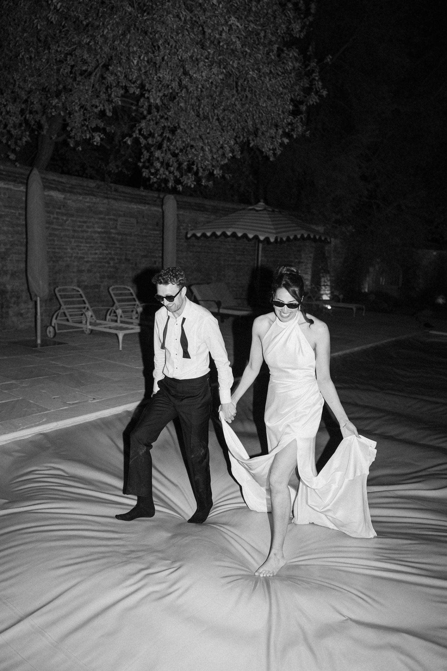 Bride and groom walk hand in hand outdoors at night wearing sunglasses in wedding attire.