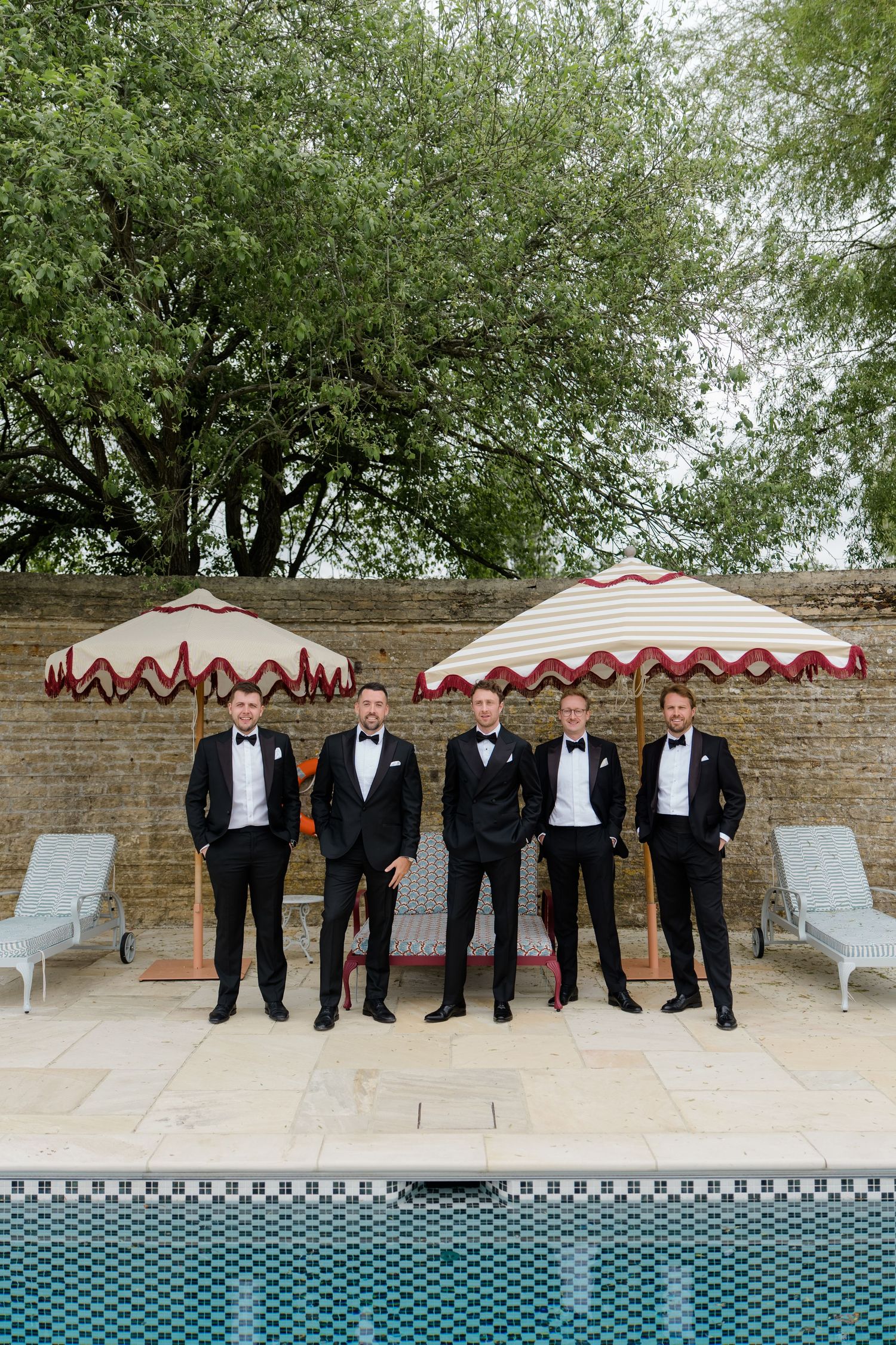 Five groomsmen in black tuxedos posing poolside with striped umbrellas and brick wall backdrop.