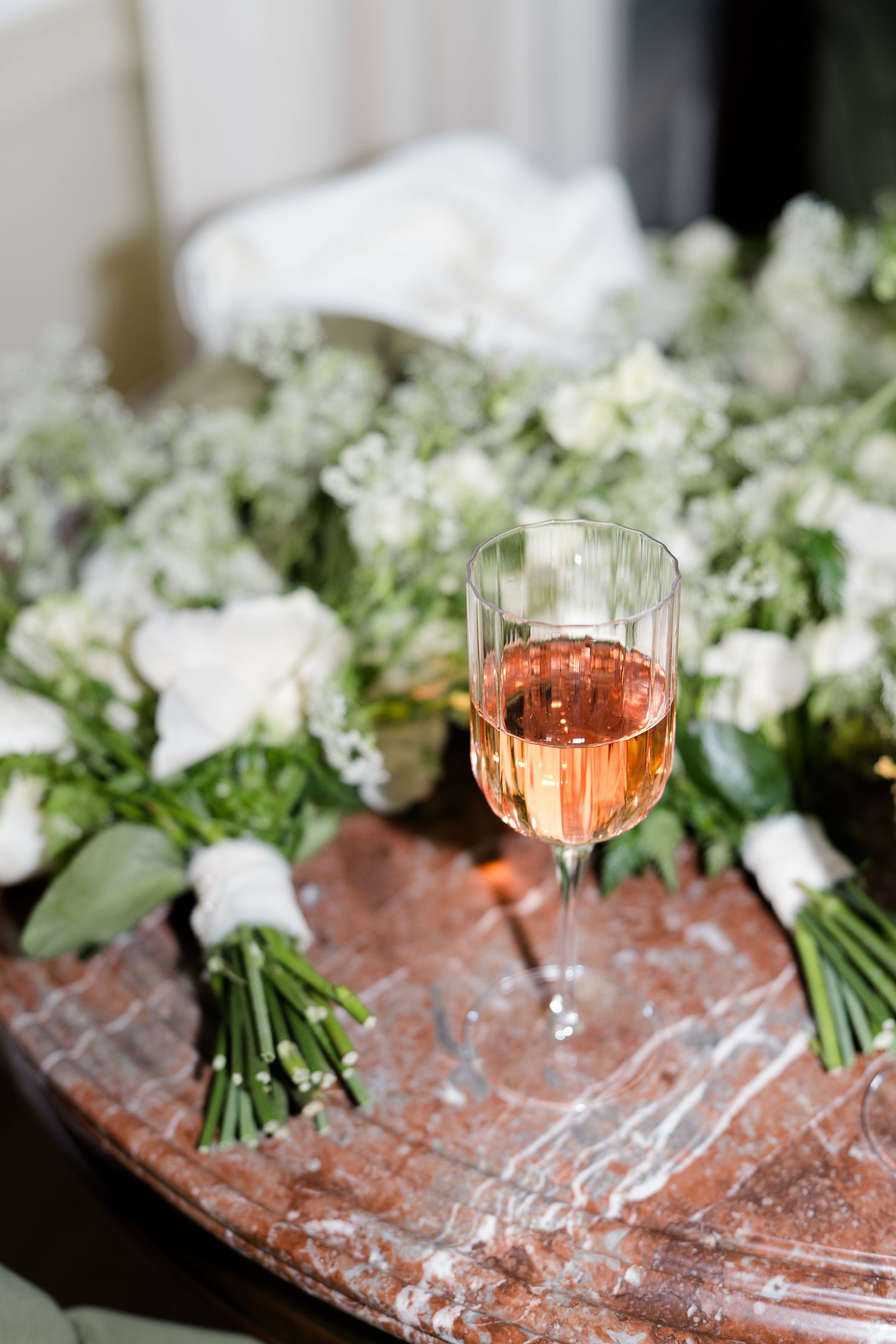 Glass of rose wine sits beside white floral wedding bouquets on a rustic wooden surface.