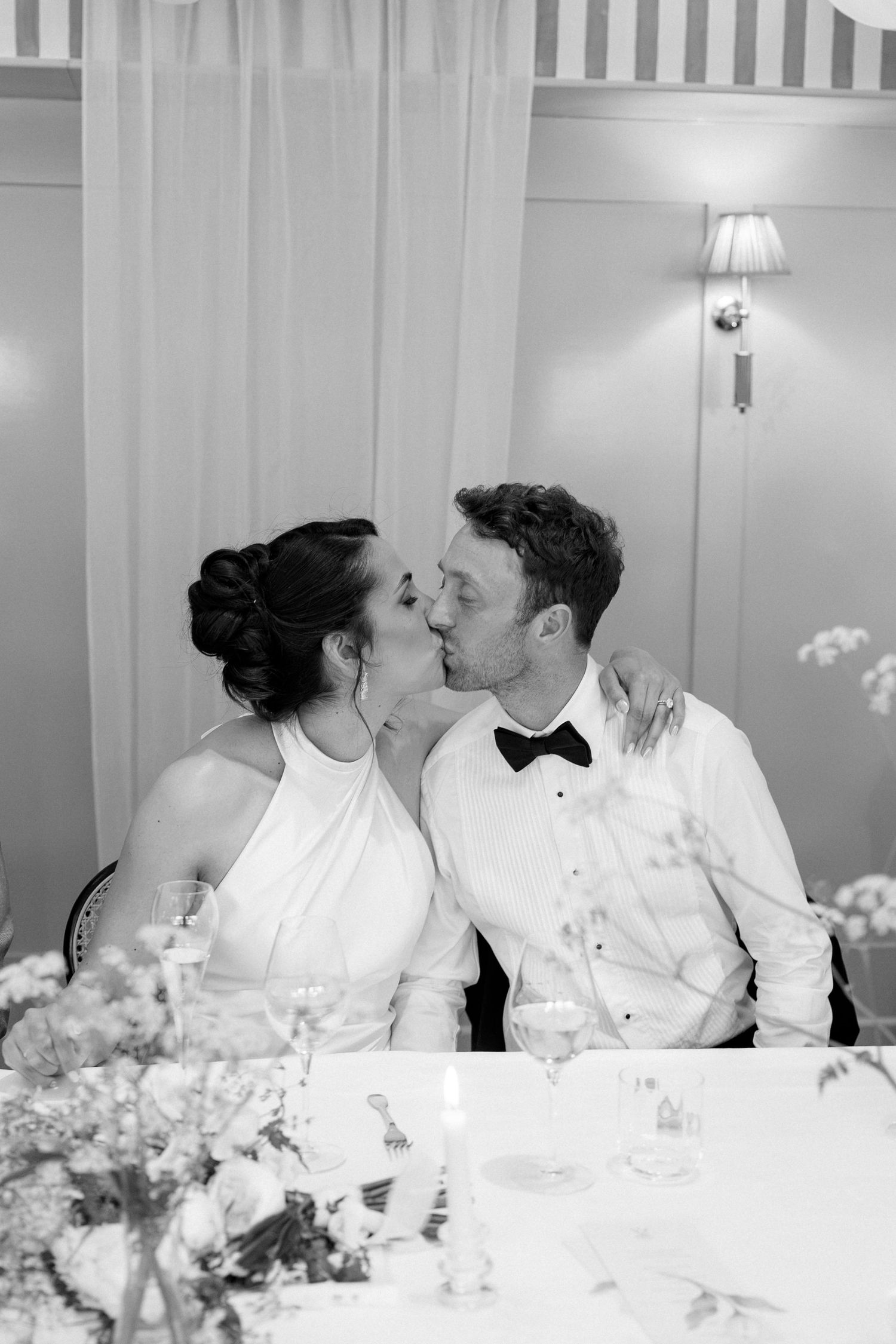 Bride and groom share a kiss at their wedding reception table in black and white photo.