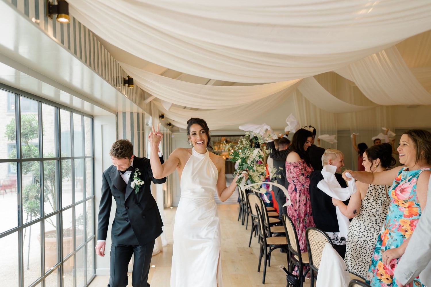 Bride in white halter gown entering wedding reception as guests cheer under white draped ceiling decor.