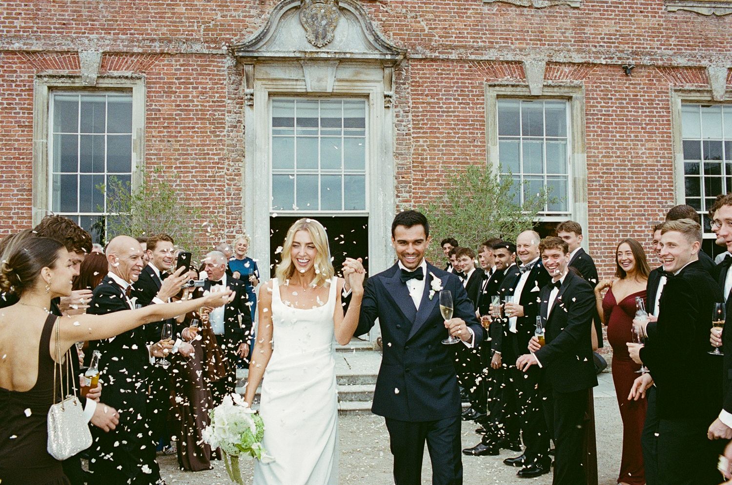 Bride and groom walk through confetti shower outside brick venue, guests celebrating joyful wedding exit.