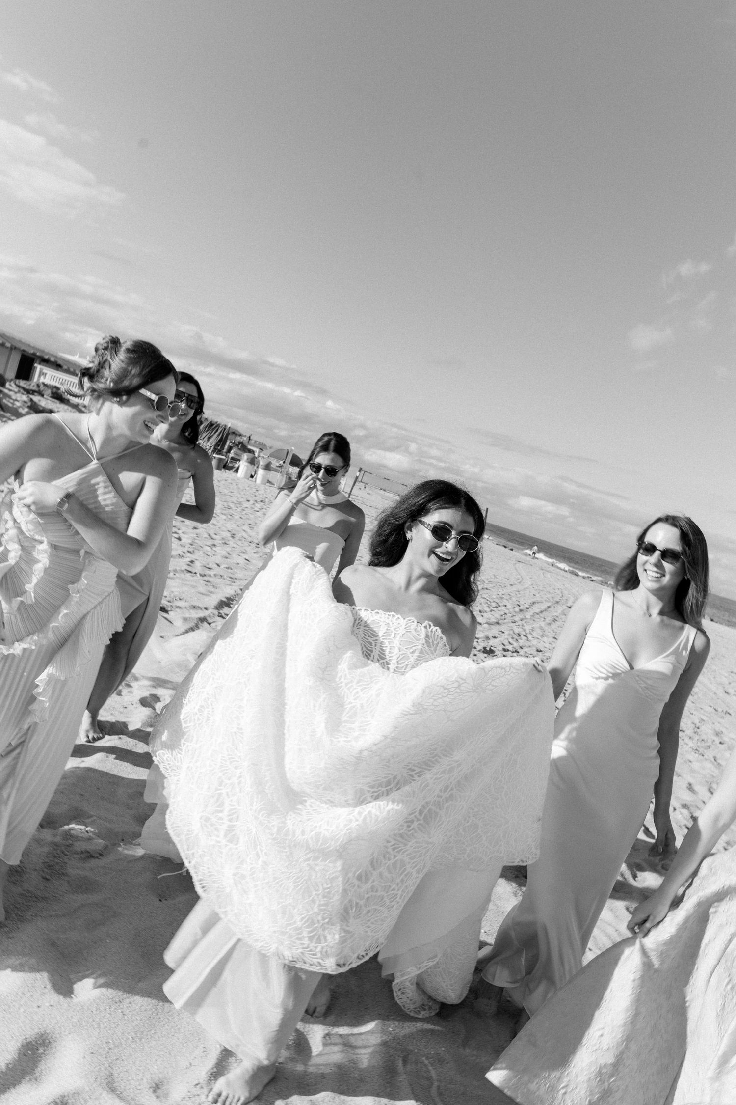 Bride and bridesmaids in white dresses laugh on a sandy beach in this black and white wedding photo.