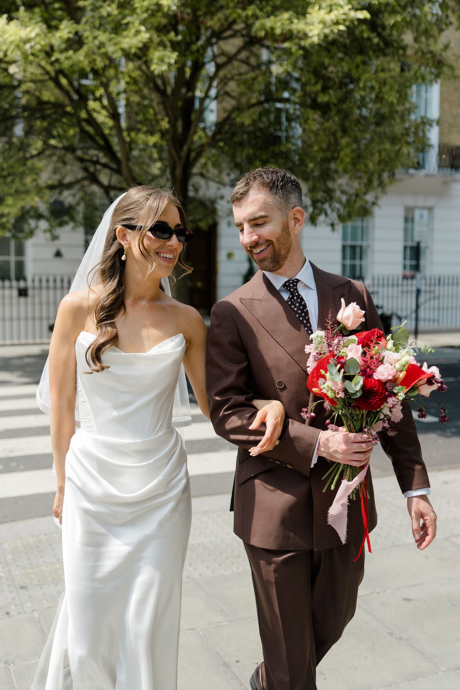 Smiling bride in white satin gown and groom in brown suit walk outdoors on sunny wedding day.