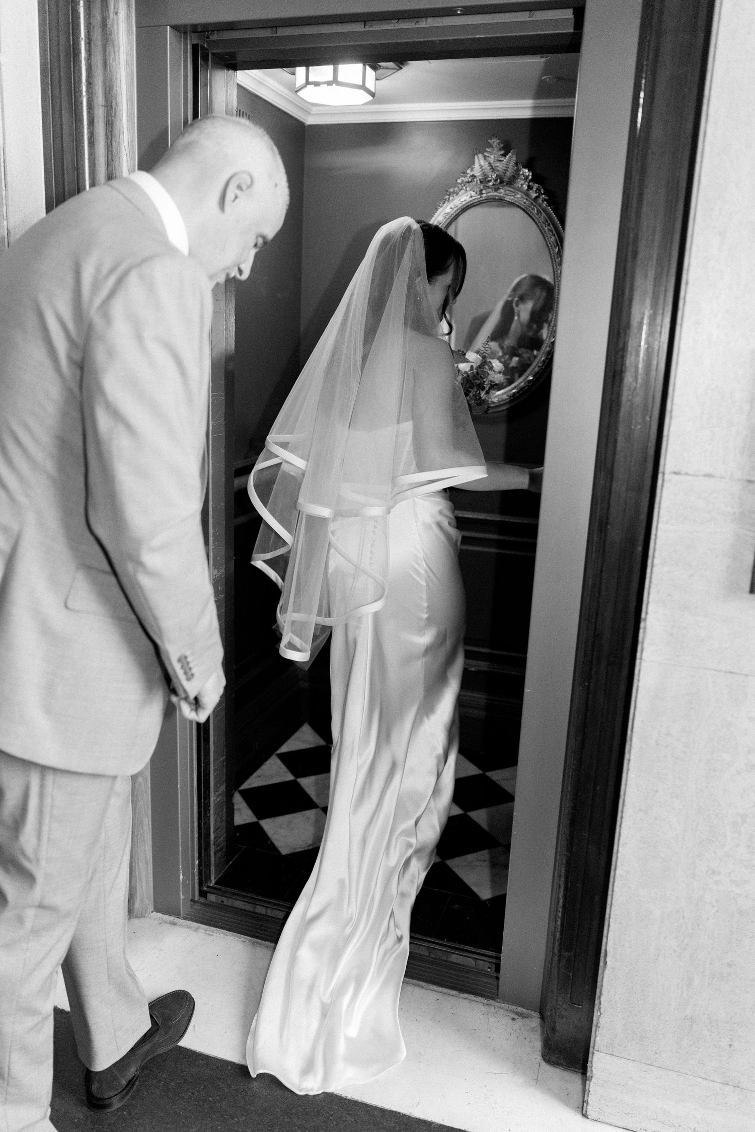 Black and white photo of bride and escort entering ornate elevator with decorative mirror.