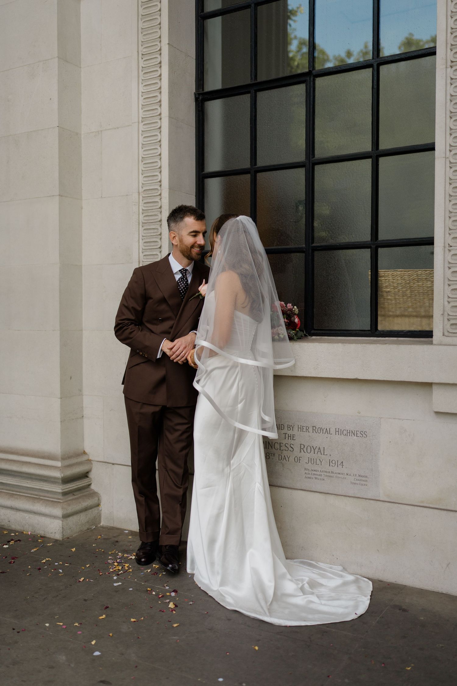 Bride in white gown and veil with groom in brown suit standing together outside a grand building.