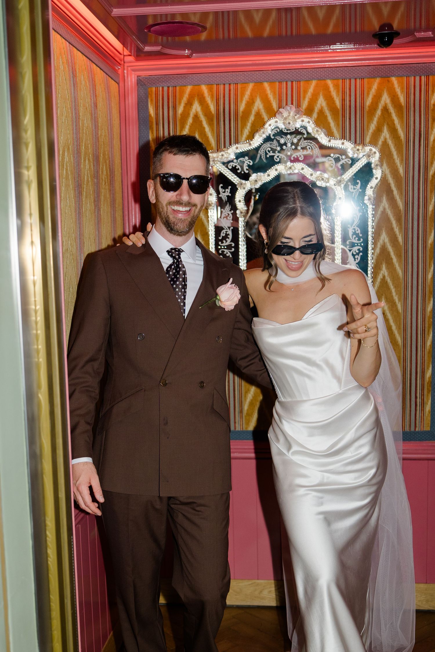 Laughing bride and groom wearing sunglasses pose together inside a colorful ornate photo booth at their wedding.