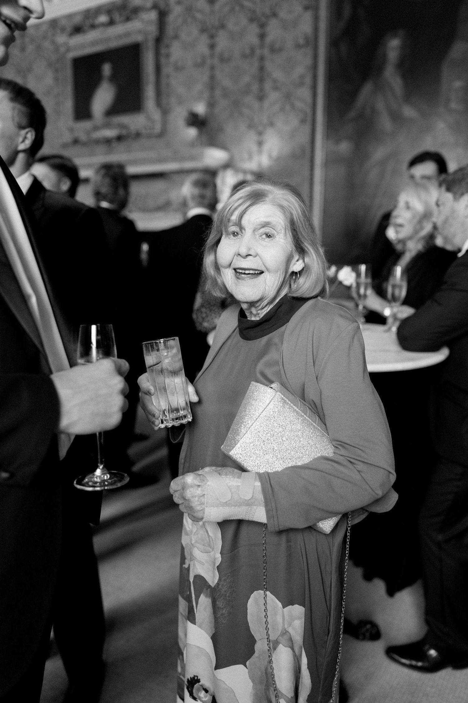 Black and white photo of elderly woman smiling at wedding reception holding a clutch purse.