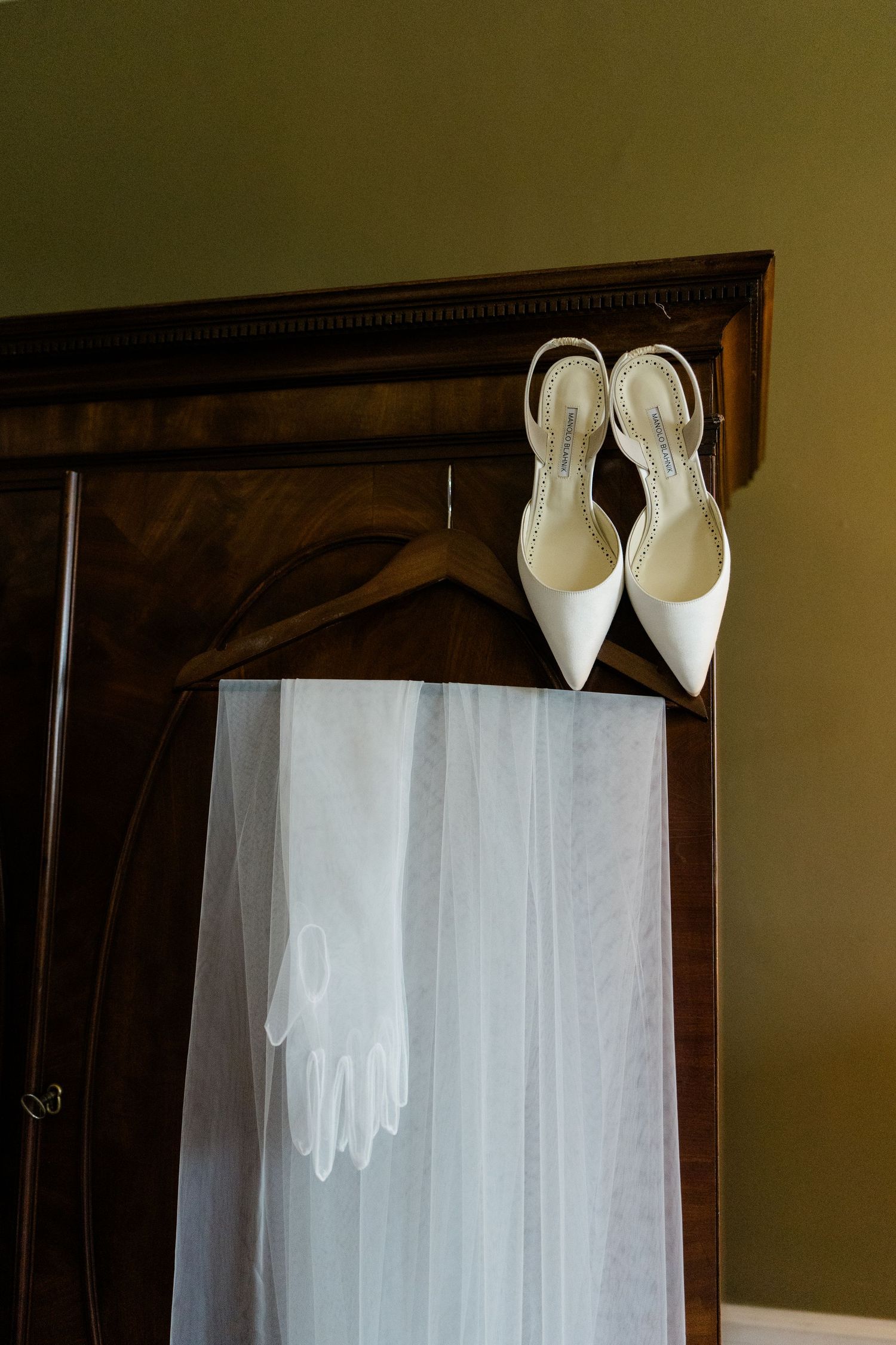 White bridal heels, lace gloves, and tulle veil displayed on a dark wooden wardrobe before a wedding.