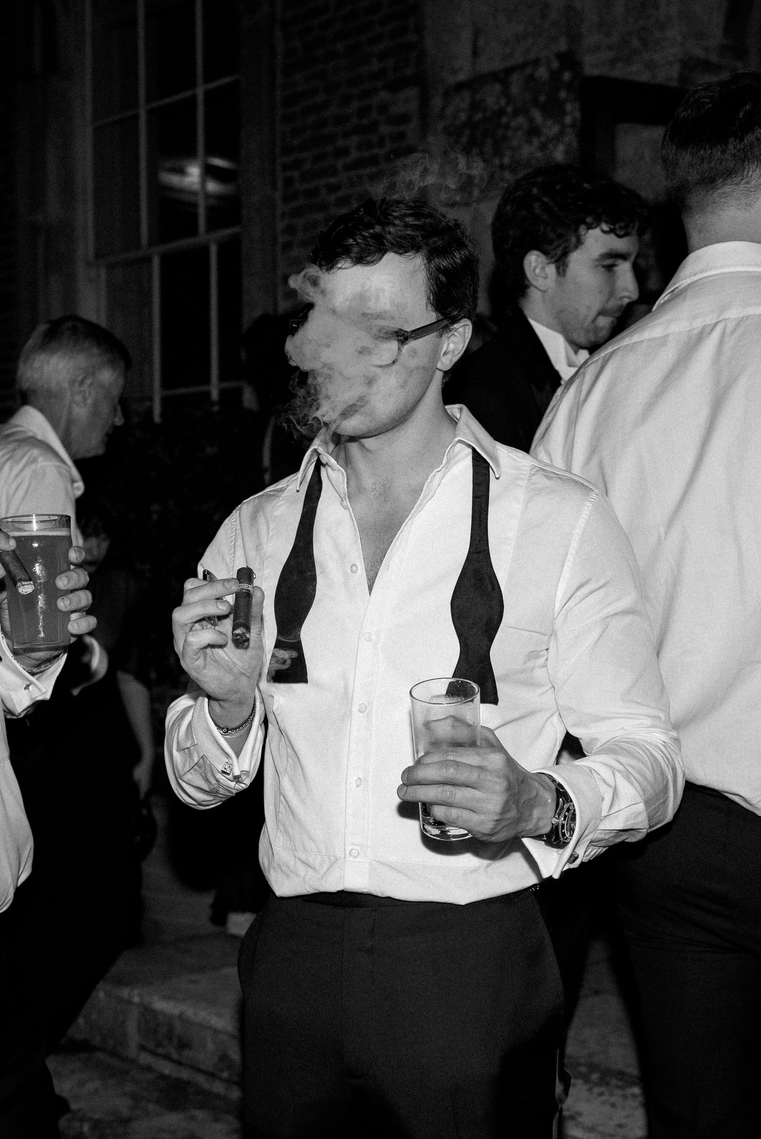 Black and white photo of a man smoking a cigar and holding drinks at a wedding party.