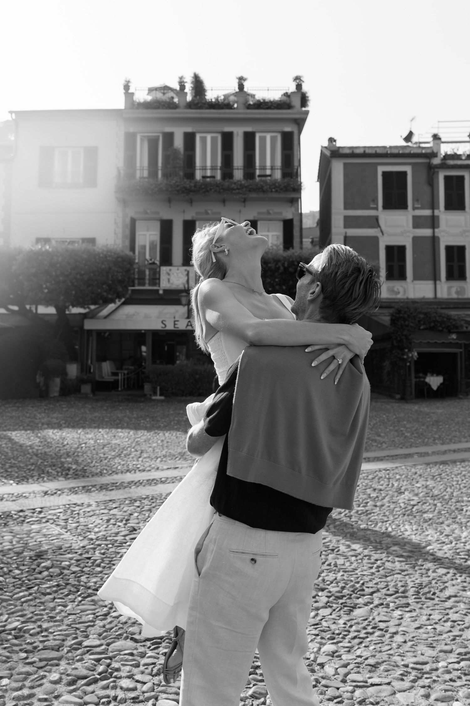 Romantic couple embracing and laughing in sunlit Italian cobblestone square, black and white portrait.