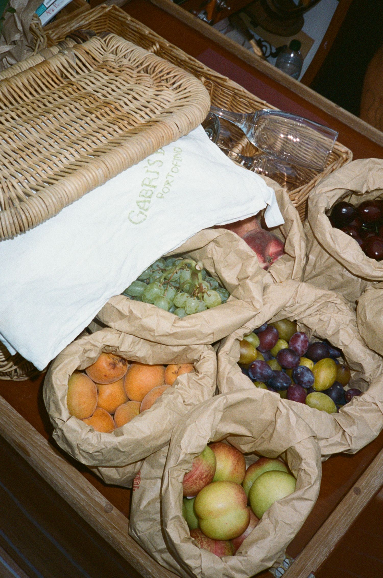 Assorted fresh fruits in paper bags arranged on wooden table with wicker basket.