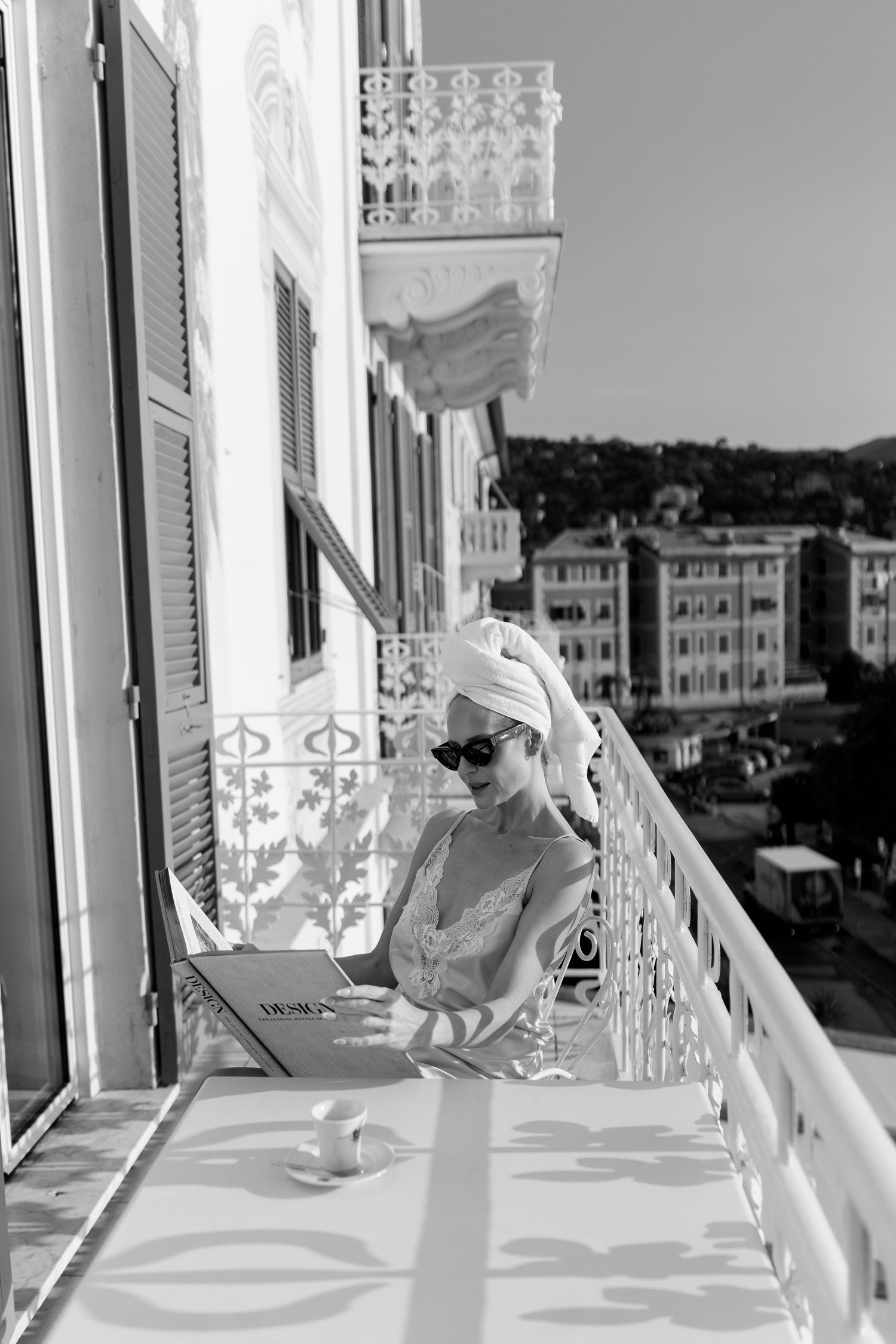 Black and white photo of woman with towel turban reading magazine on ornate hotel balcony.