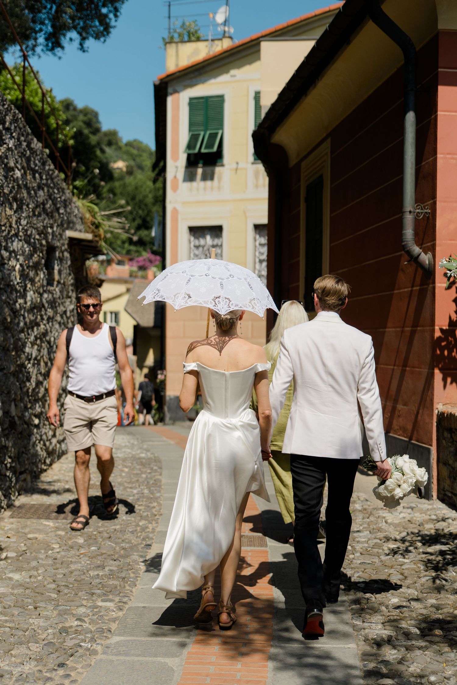 Bride in a white off-shoulder gown holding a lace parasol walks with groom through a narrow Italian village street.