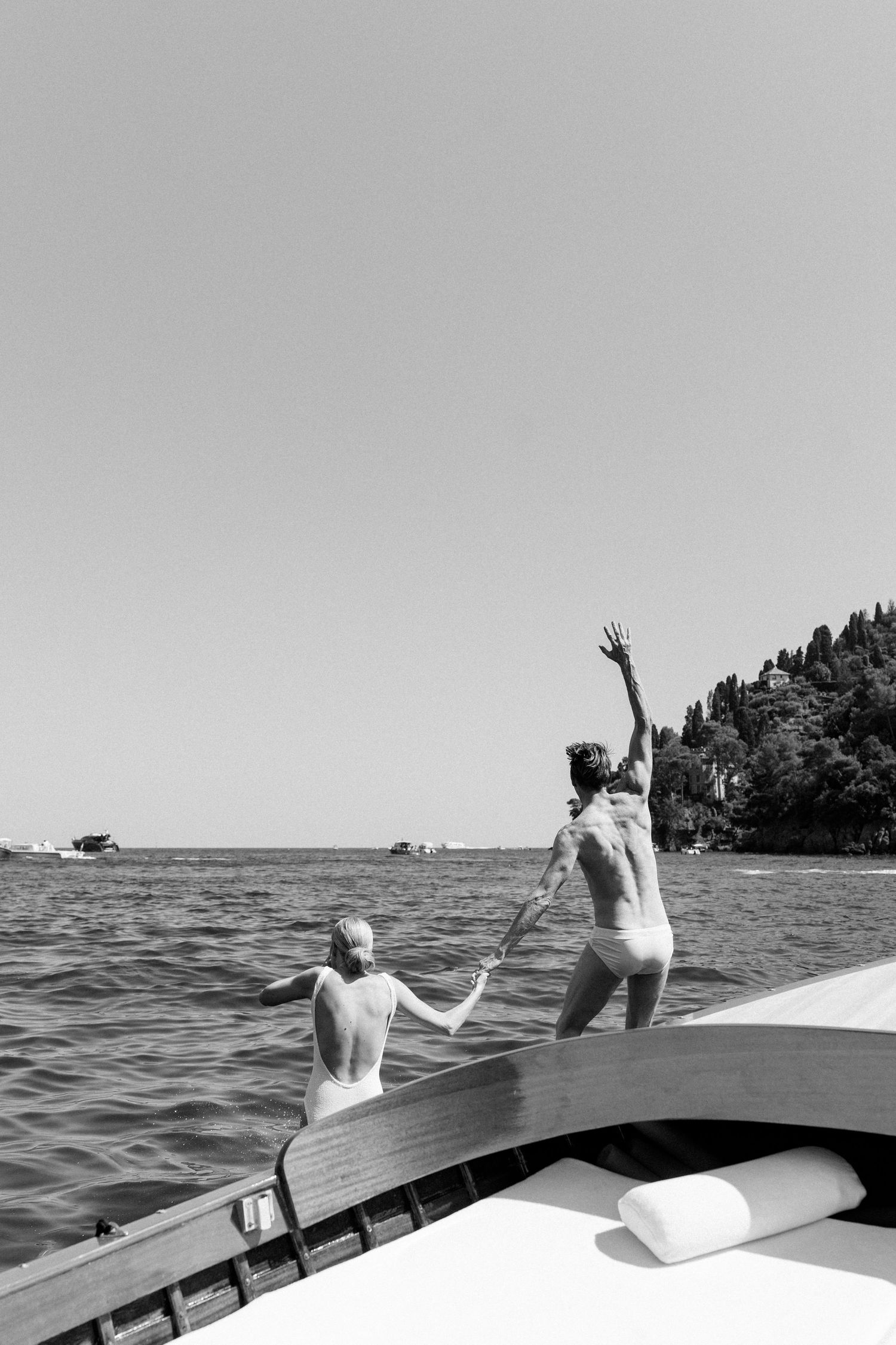 Two people jumping off the bow of a wooden boat into calm open waters on a sunny day.