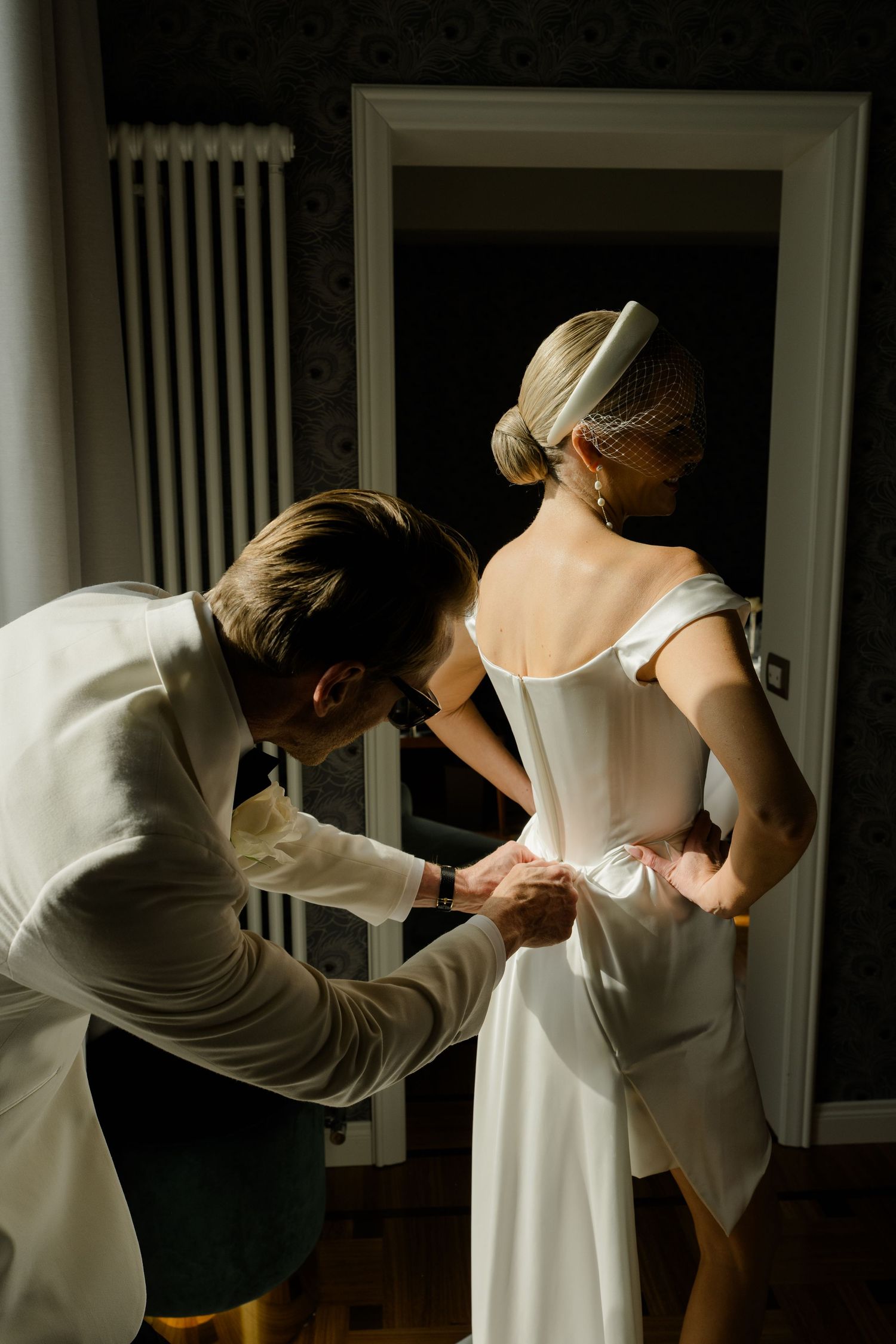 Groom carefully fastens bride's white wedding dress in elegant dimly lit hotel room.