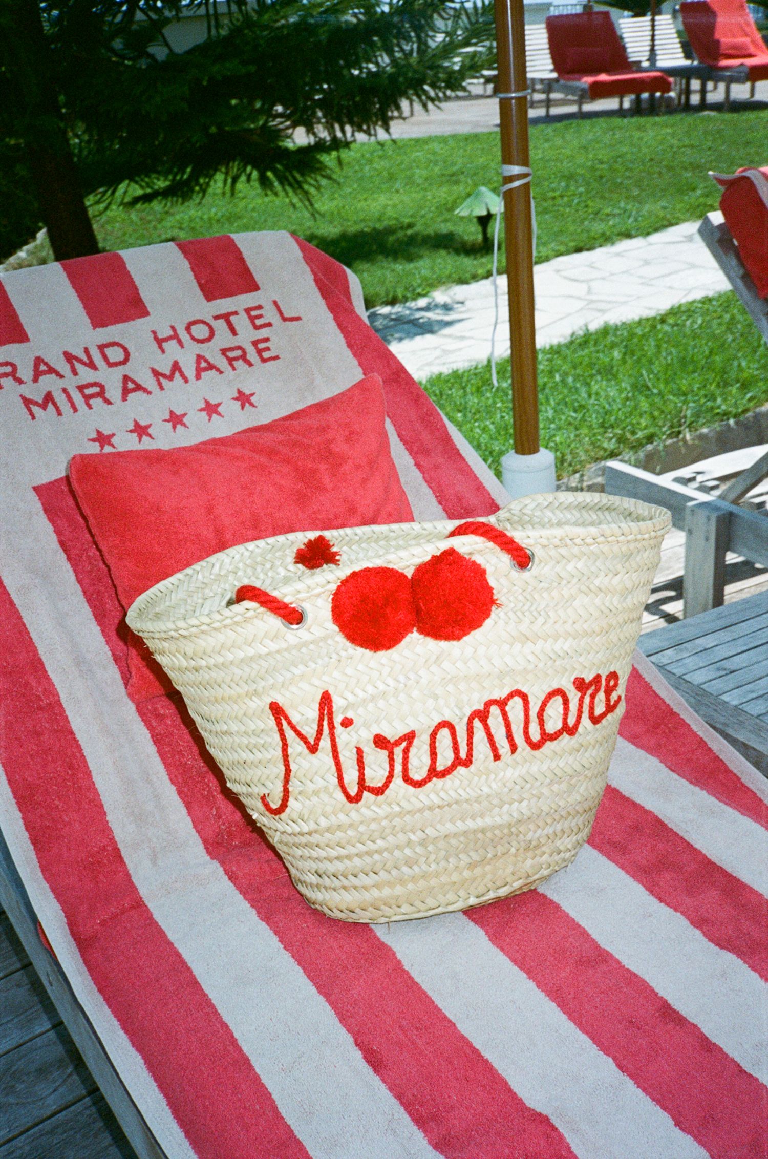 A woven straw tote bag with red Miramare lettering on a striped red and white Grand Hotel Miramare beach towel.