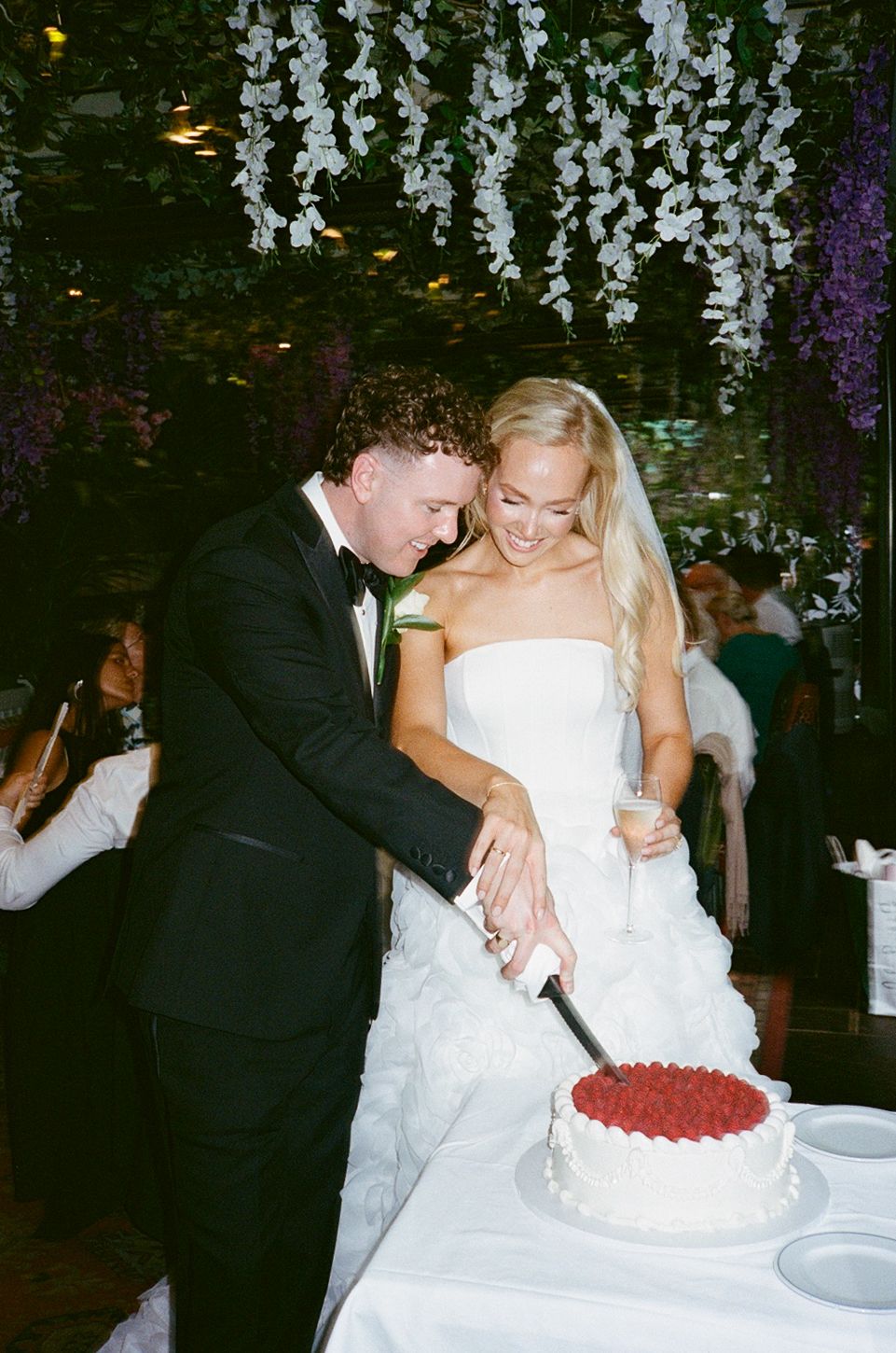 Bride and groom cutting white wedding cake topped with raspberries under floral canopy at elegant reception.
