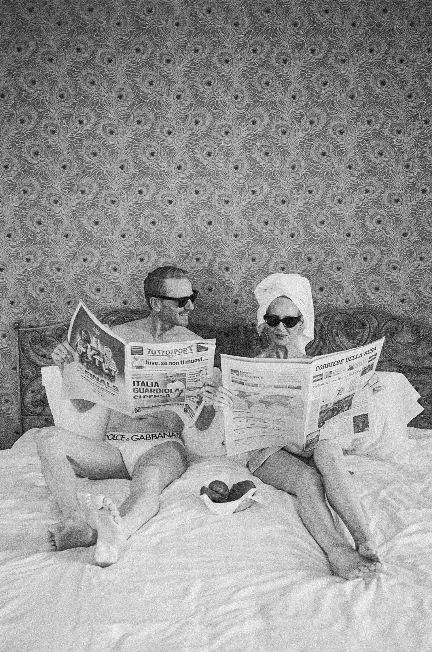 Black and white photo of couple in sunglasses reading Italian newspapers on bed with ornate wallpaper.