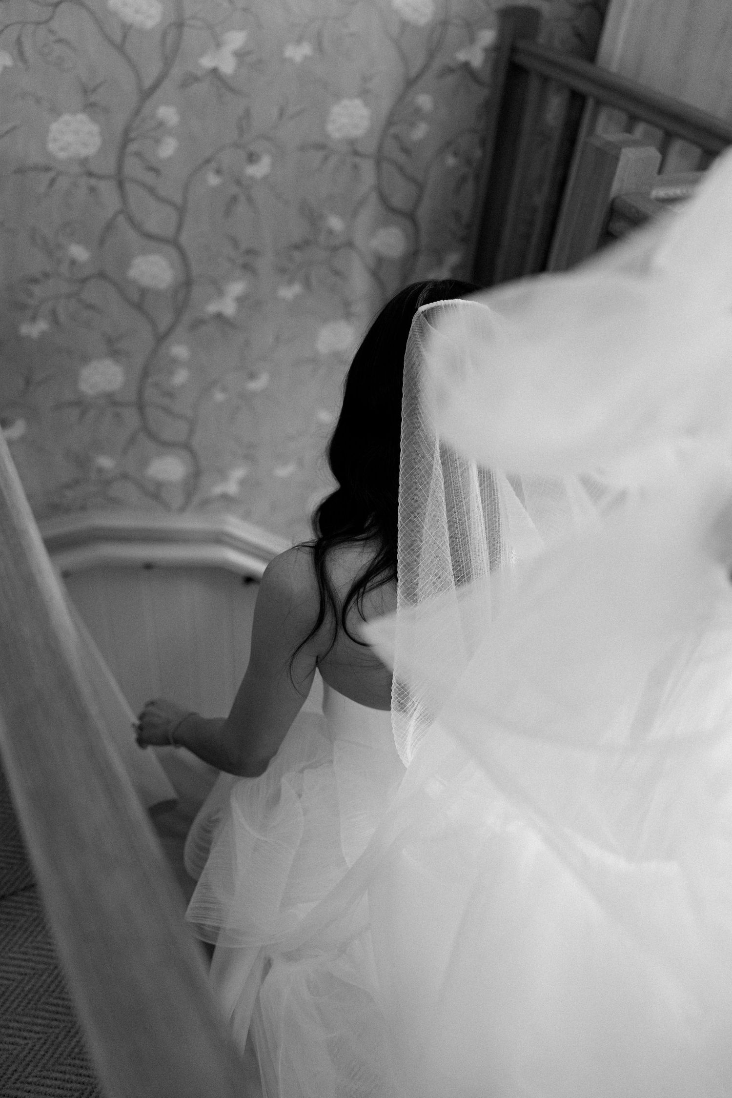 A bride in a flowing white wedding dress descending stairs, captured in artistic black and white.