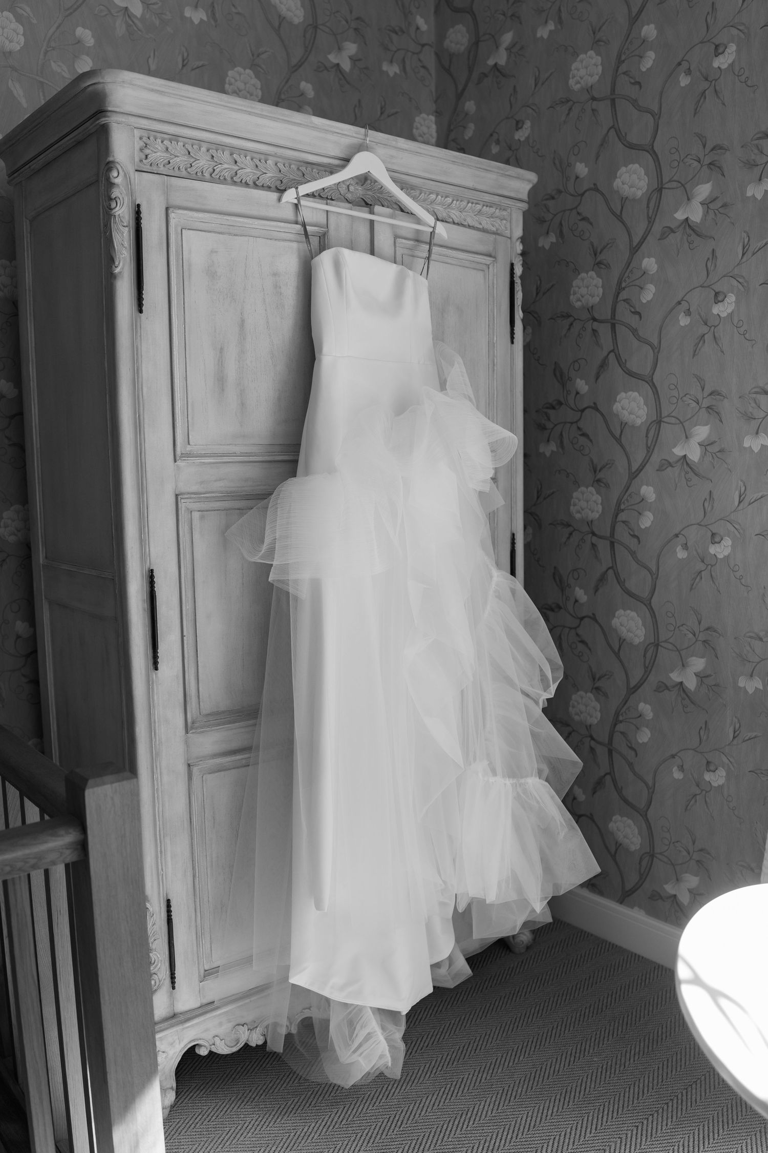 White wedding dress hanging on vintage armoire against floral wallpaper in black and white.