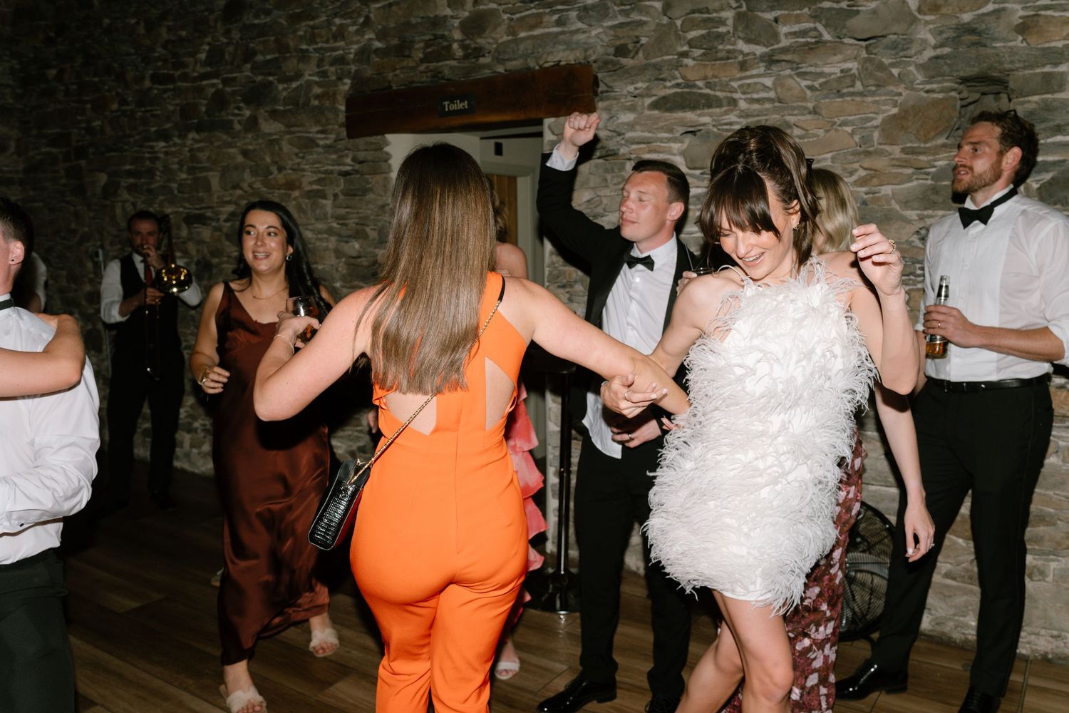 Wedding party guests dancing energetically in a rustic stone-walled venue during a celebration.