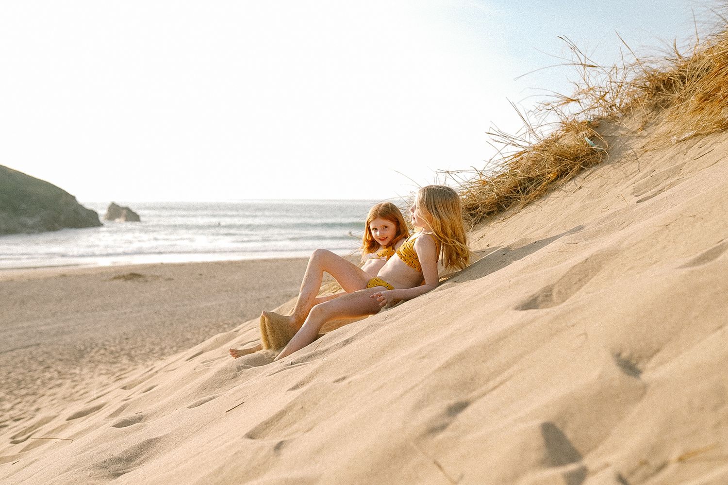 People sliding down sandy beach dunes at sunset with ocean in background.