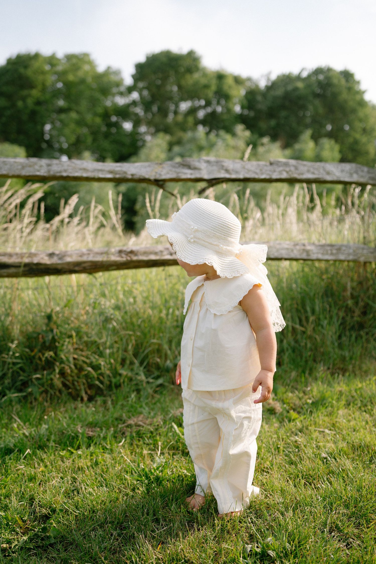 A small figure in white clothing walks through tall grass near a wooden fence.