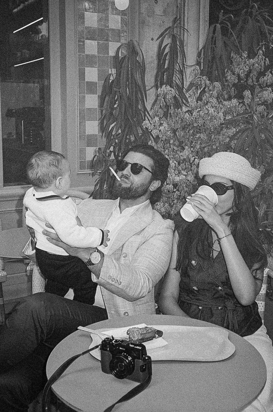 Family at cafe table with baby, man in sunglasses and woman drinking coffee, camera on table.