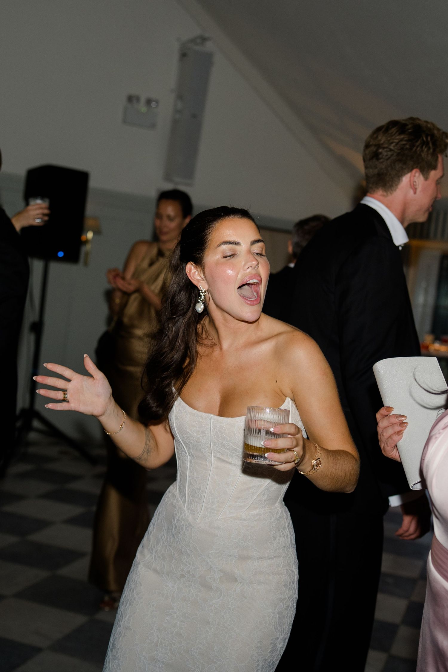 Joyful bride in strapless lace wedding gown dancing and singing with drink in hand at wedding reception.