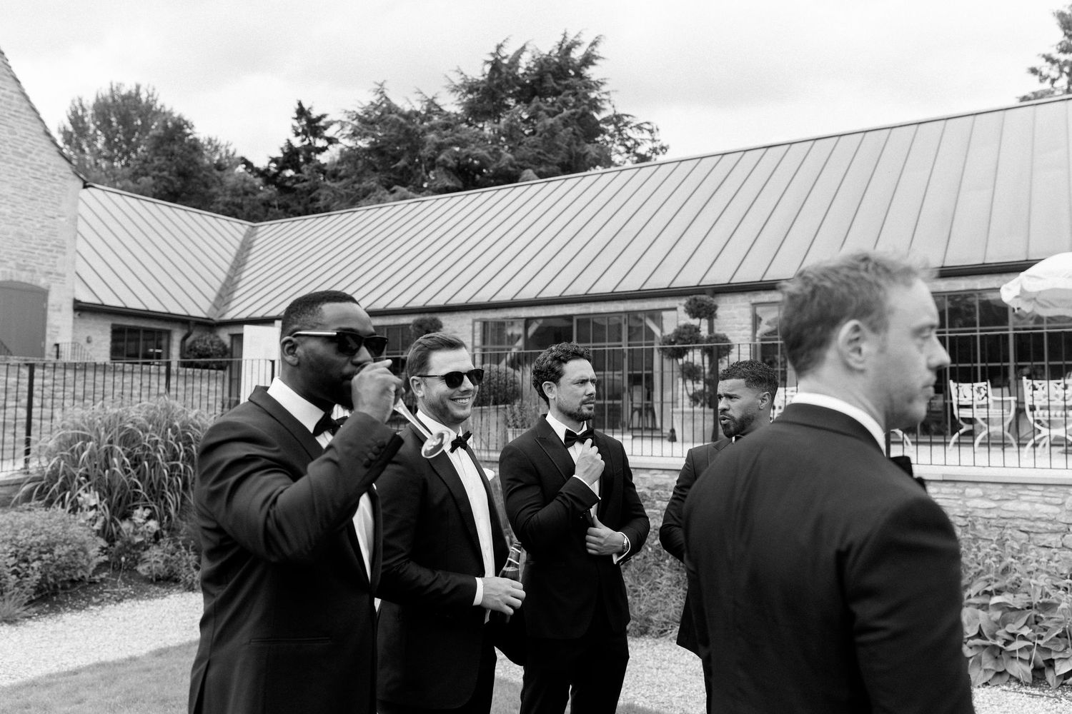 A black and white photo of four groomsmen in tuxedos gathered outside a wedding venue building.