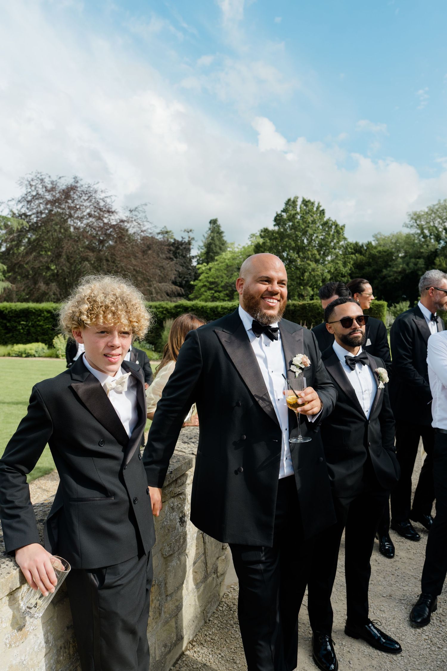 Smiling bald groom with boutonniere stands with curly-haired boy and groomsmen at outdoor wedding.