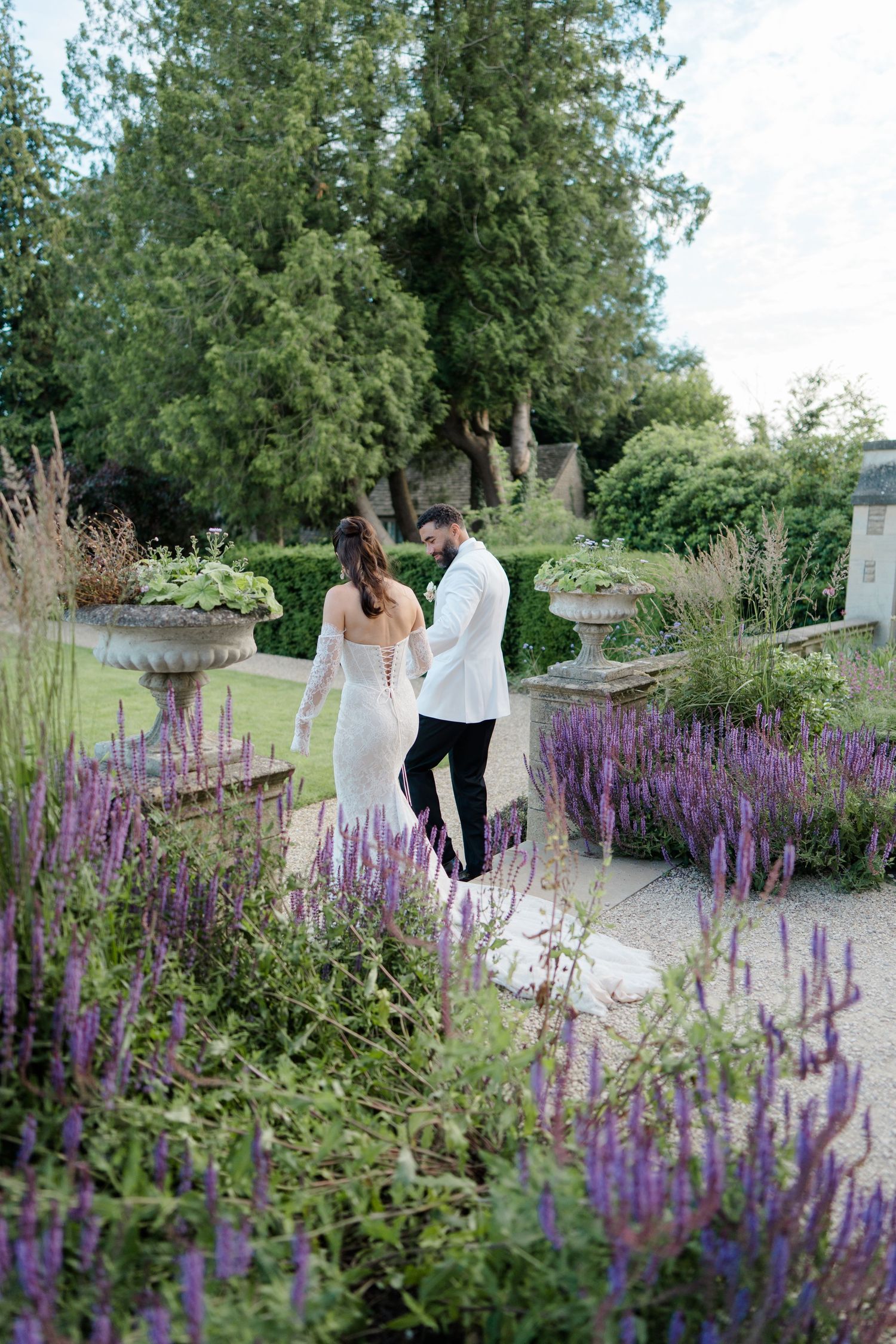 Bride and groom walk hand in hand through lush garden path lined with purple lavender flowers.